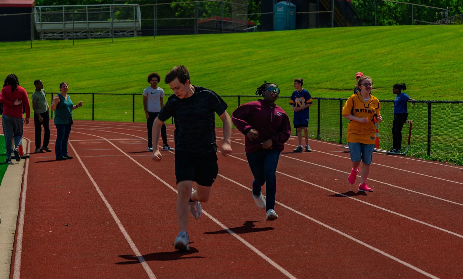 Students racing on a red track during a school field day event with spectators watching on a sunny day.