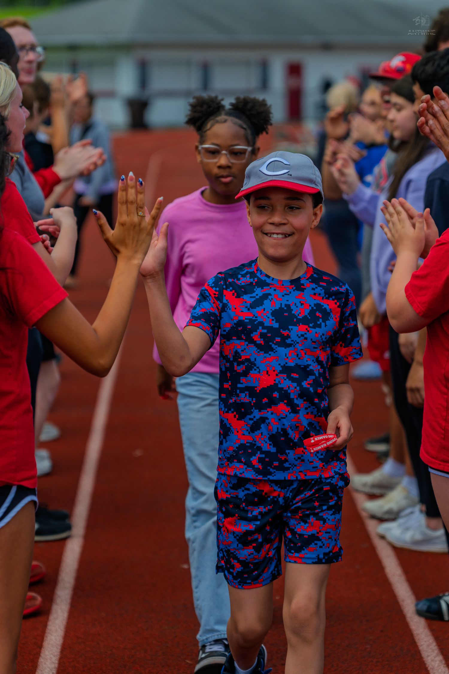 A smiling boy in a blue camo outfit high-fives peers while walking through a celebratory line on a track.