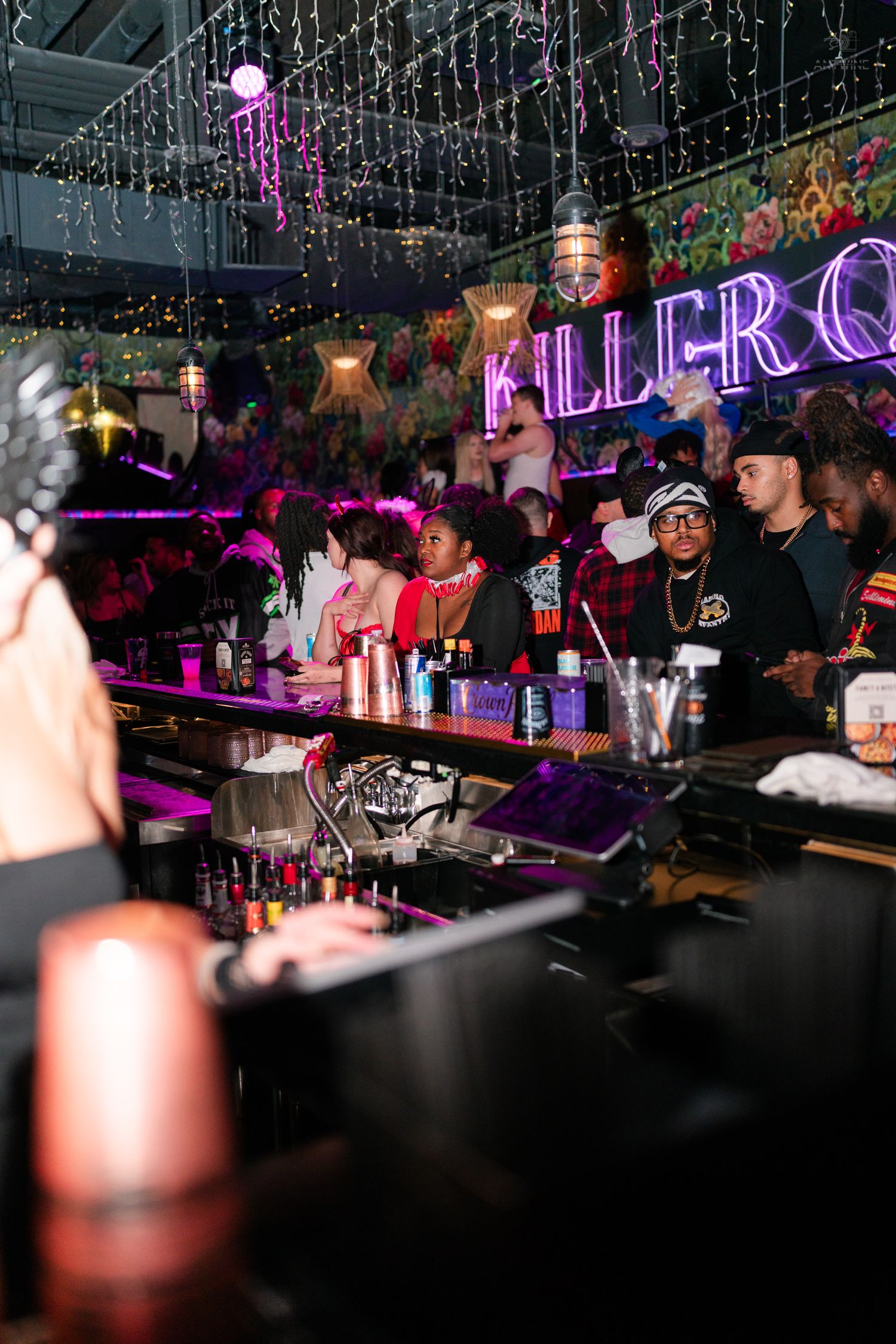 Bartender and patrons at a colorfully decorated bar with a Killer Queen neon sign.