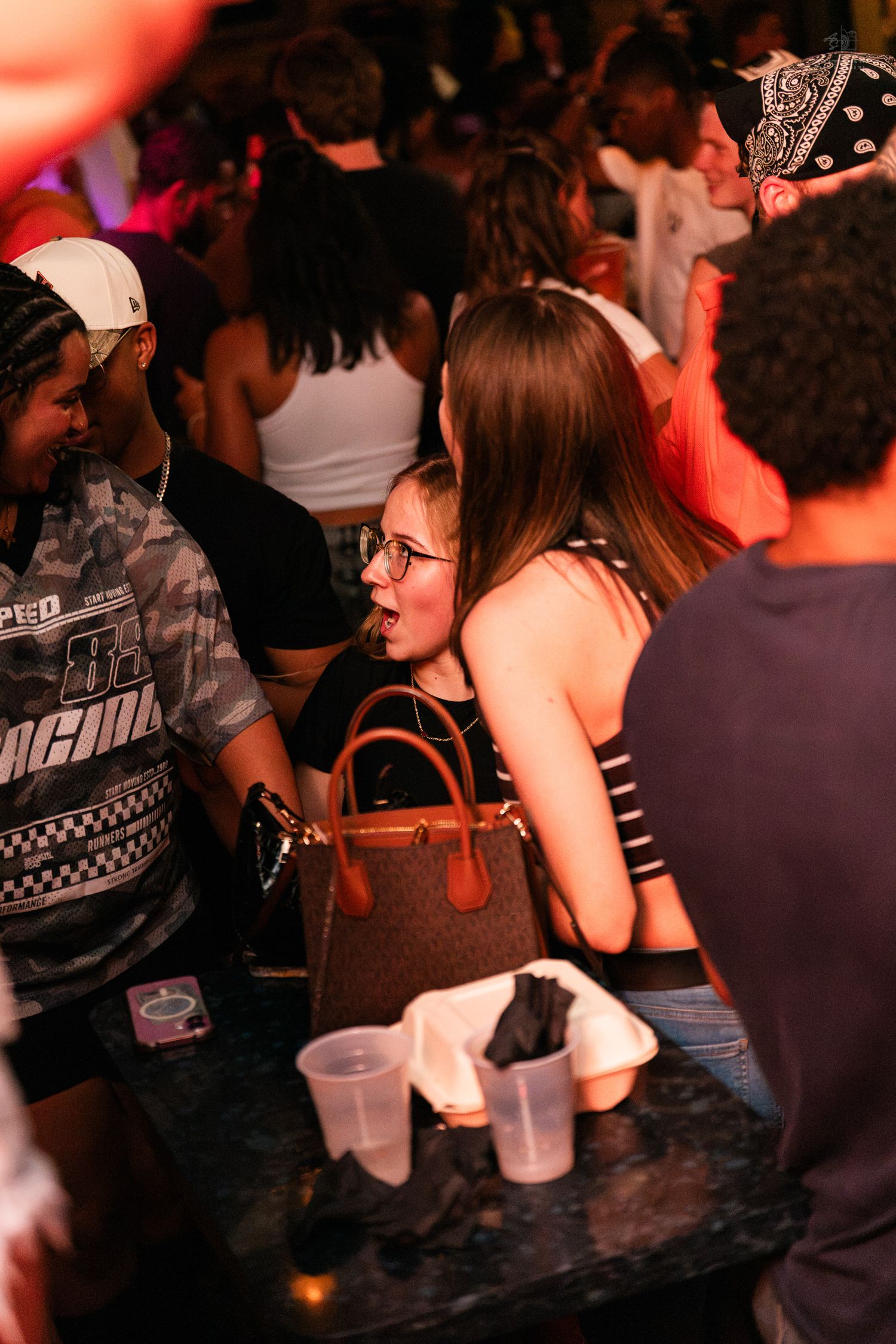 Young woman with glasses and handbag socializing at a crowded nightclub event.
