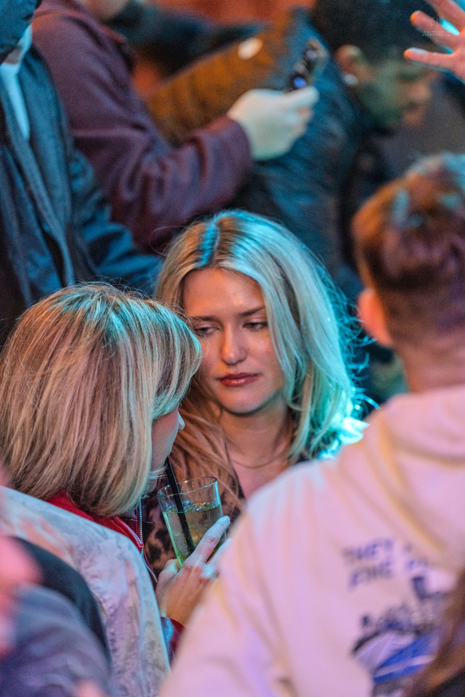 Two blonde women talking closely together at a crowded nightclub or bar event.