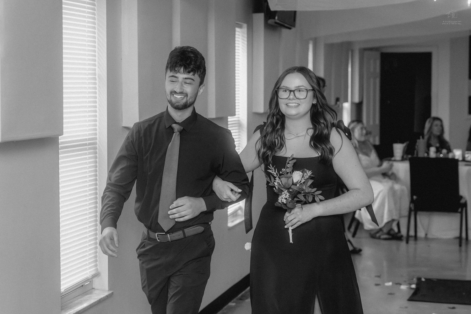 A couple in formal attire walks arm-in-arm at a wedding reception in a black and white photo.