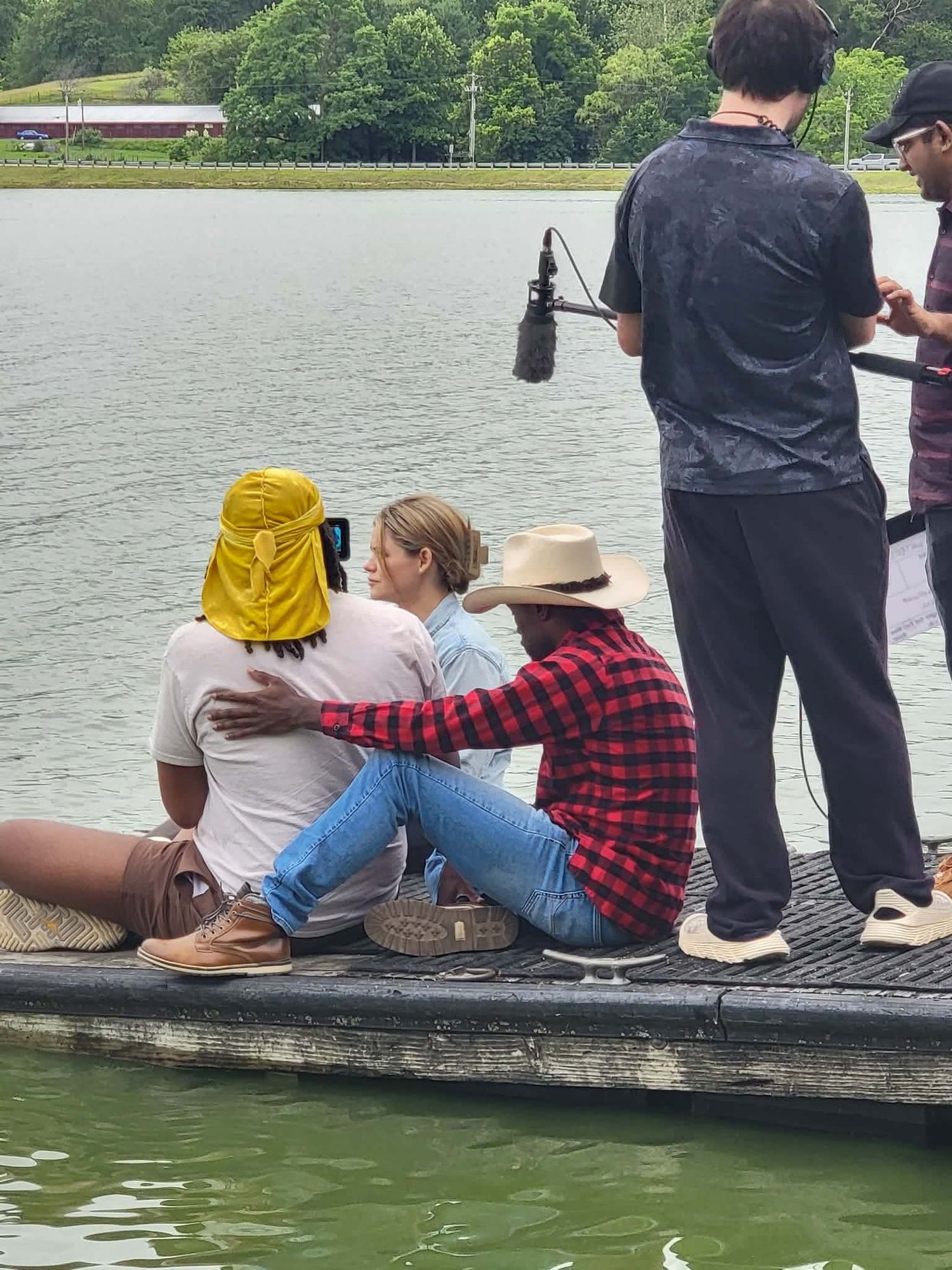 Three people sit together on a lakeside dock while a cameraman films with a boom mic nearby.