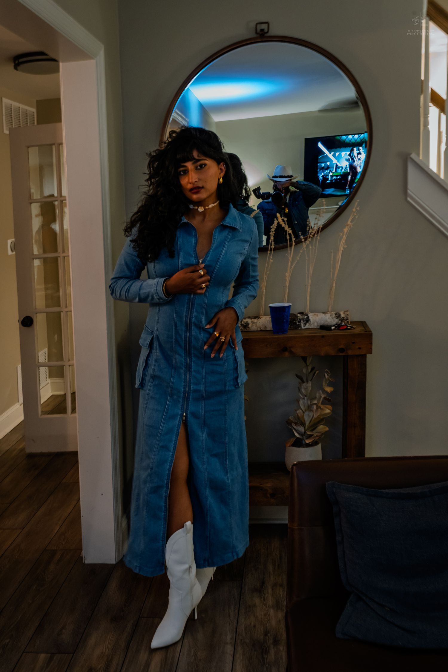 Stylish woman in blue denim maxi dress with white cowboy boots posing by round mirror indoors.