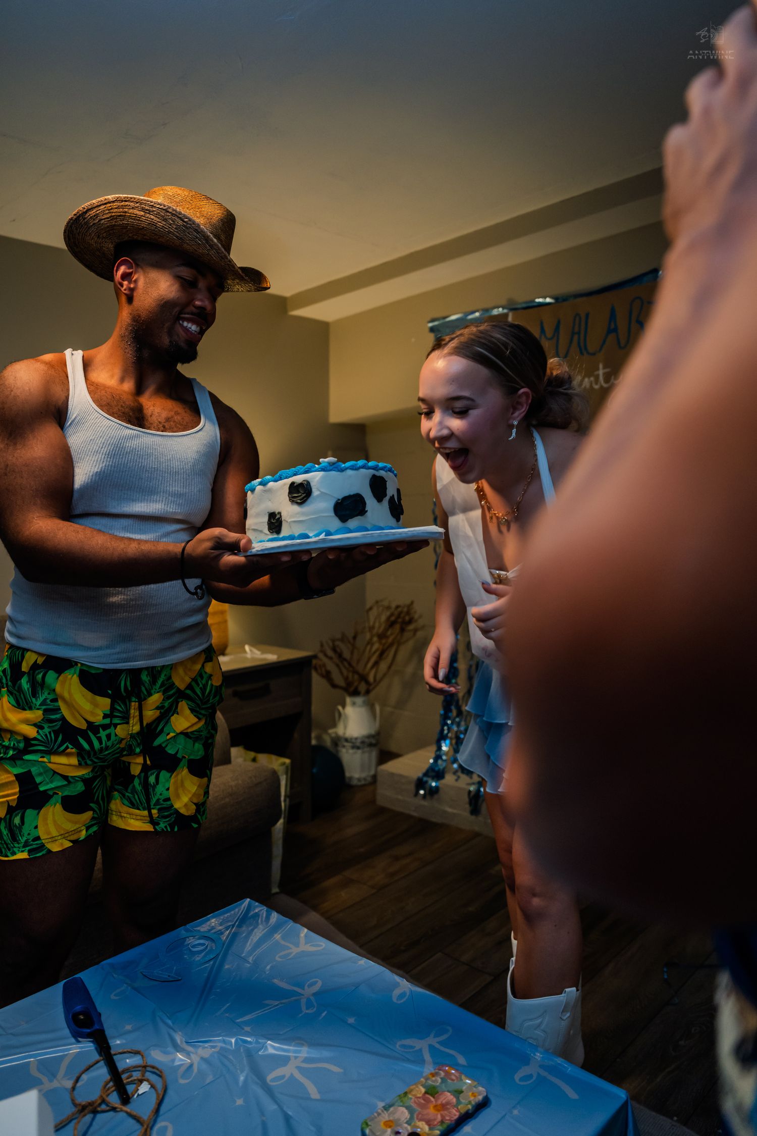 Man in straw hat and tropical shorts presents birthday cake to smiling woman at indoor party.