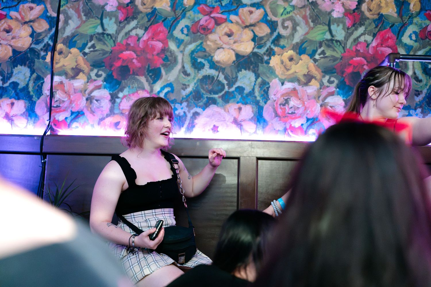 Young woman with pink hair laughing and holding a microphone at a vibrant nightclub with floral wallpaper.