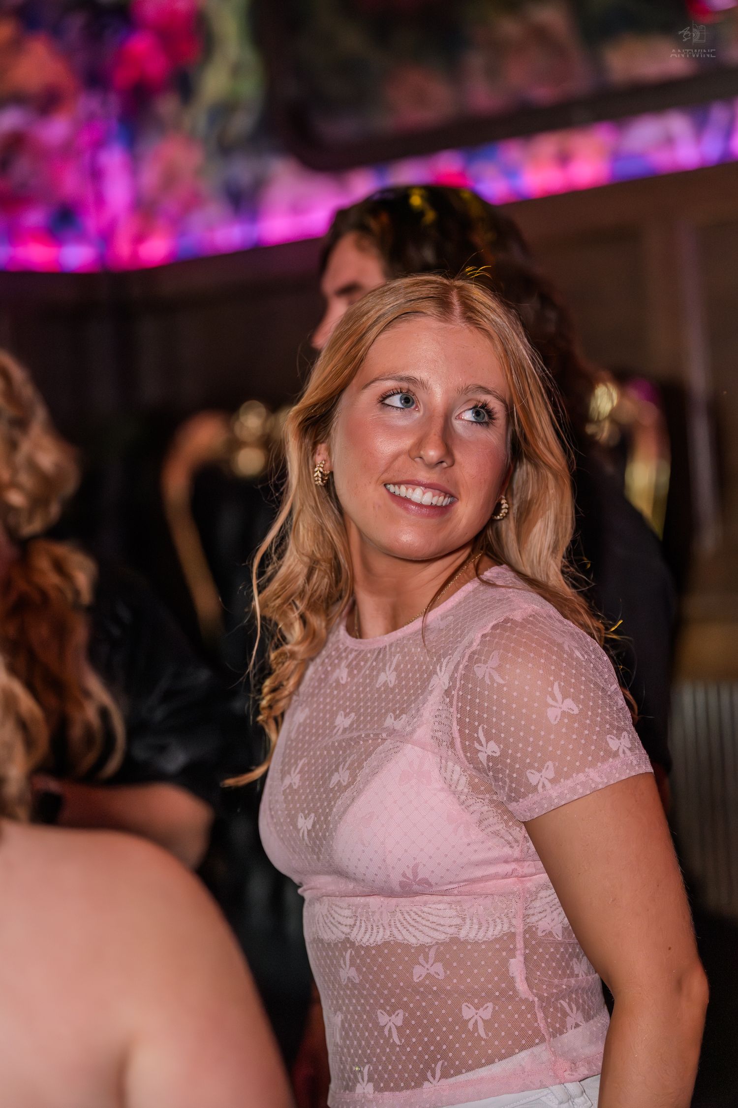 Smiling blonde woman in sheer pink top enjoying a vibrant nightclub event.