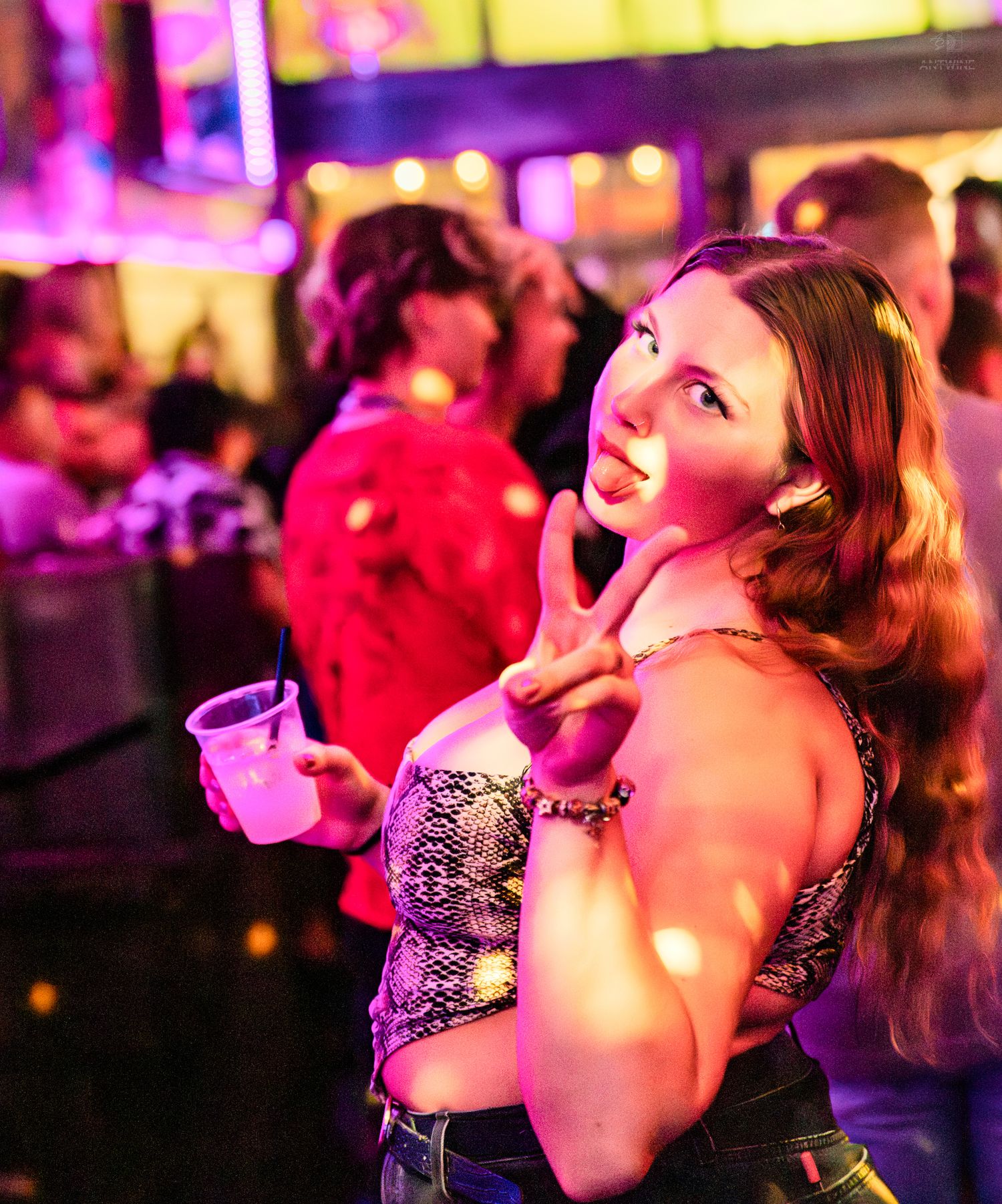 Smiling woman in snakeskin top holding drink and making peace sign at nightclub.