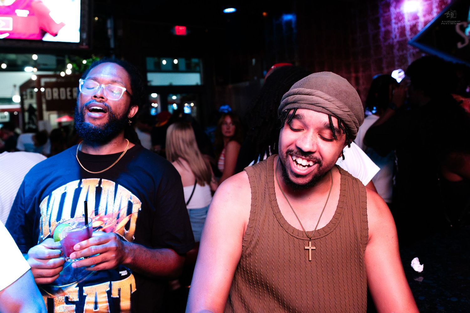 Two men smiling and enjoying drinks at a lively nightclub party in Miami Heat gear.