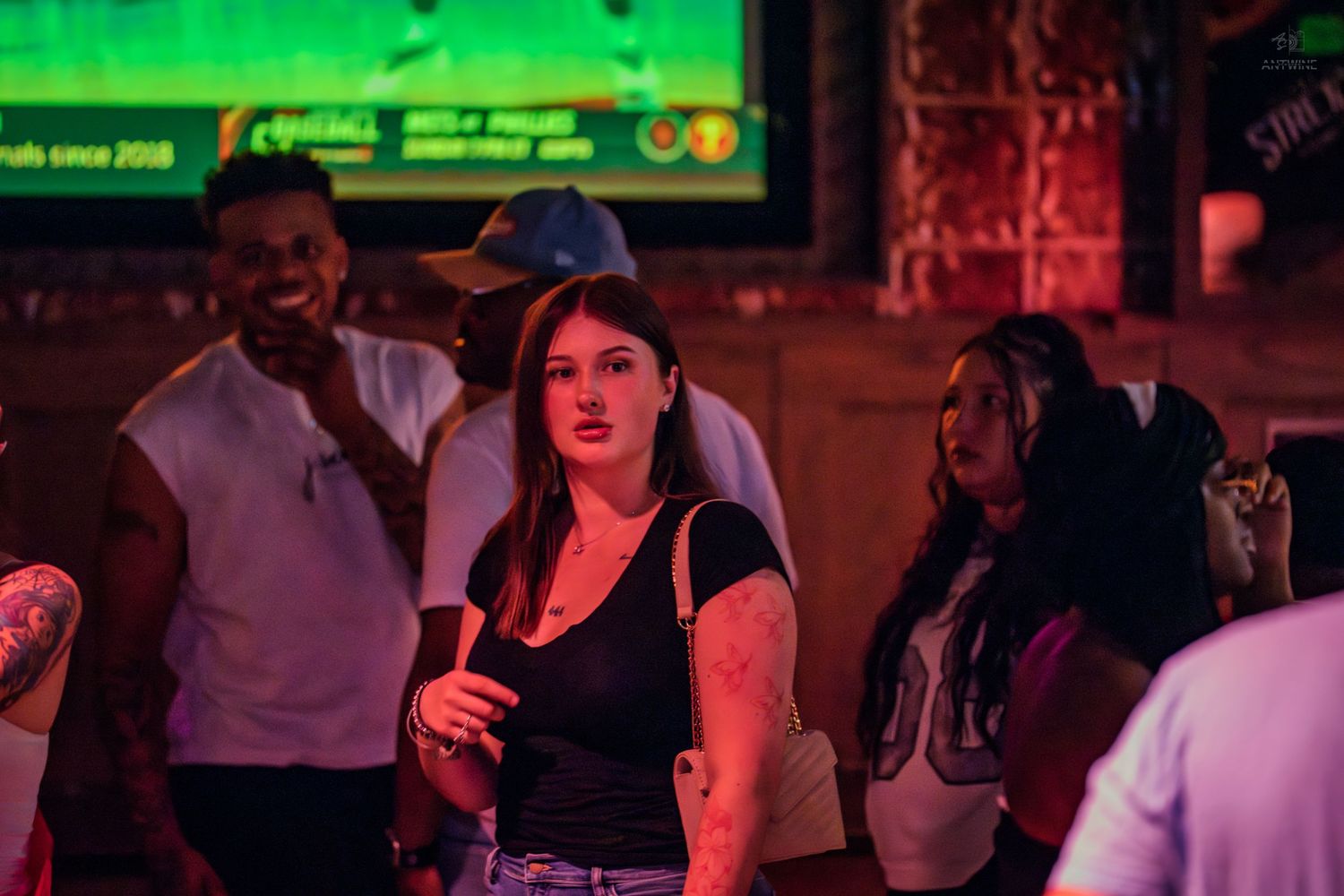 Young woman in black tank top holding drink at vibrant neon-lit bar with crowd.