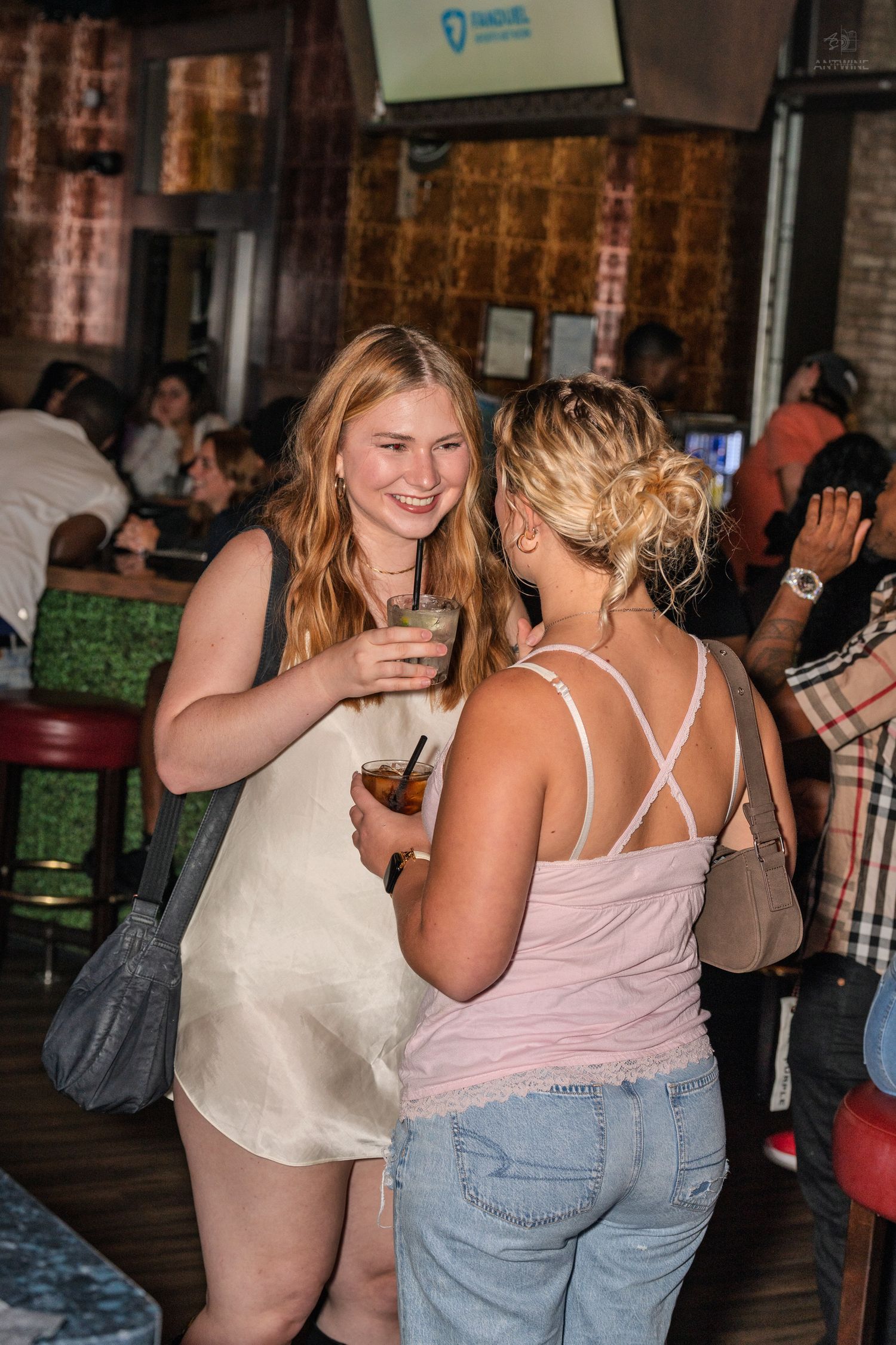 Two women socializing at a busy bar, blonde in white dress holding cocktail, other in pink top.