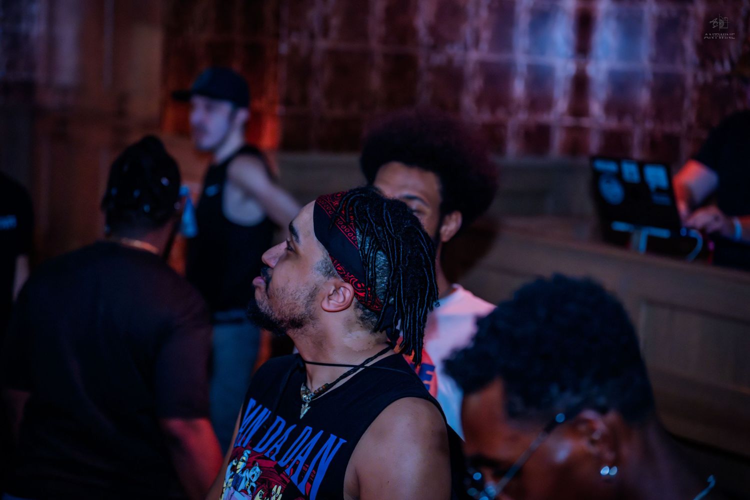 A man with braided hair looks upward in a dark nightclub surrounded by other guests.
