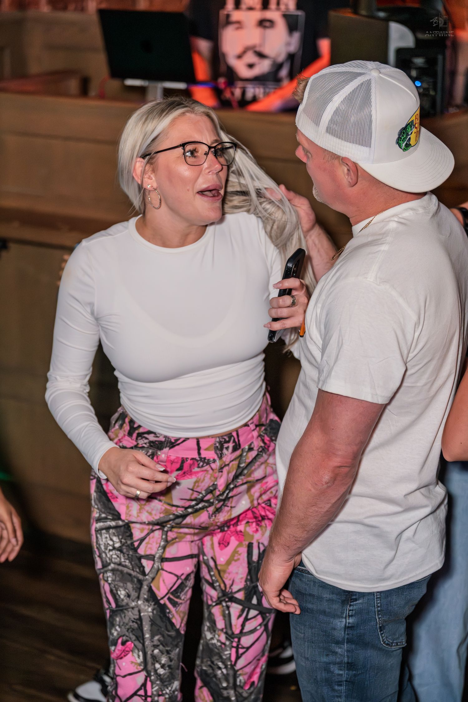 Blonde woman in white top and pink camo pants laughing with a man at a bar.