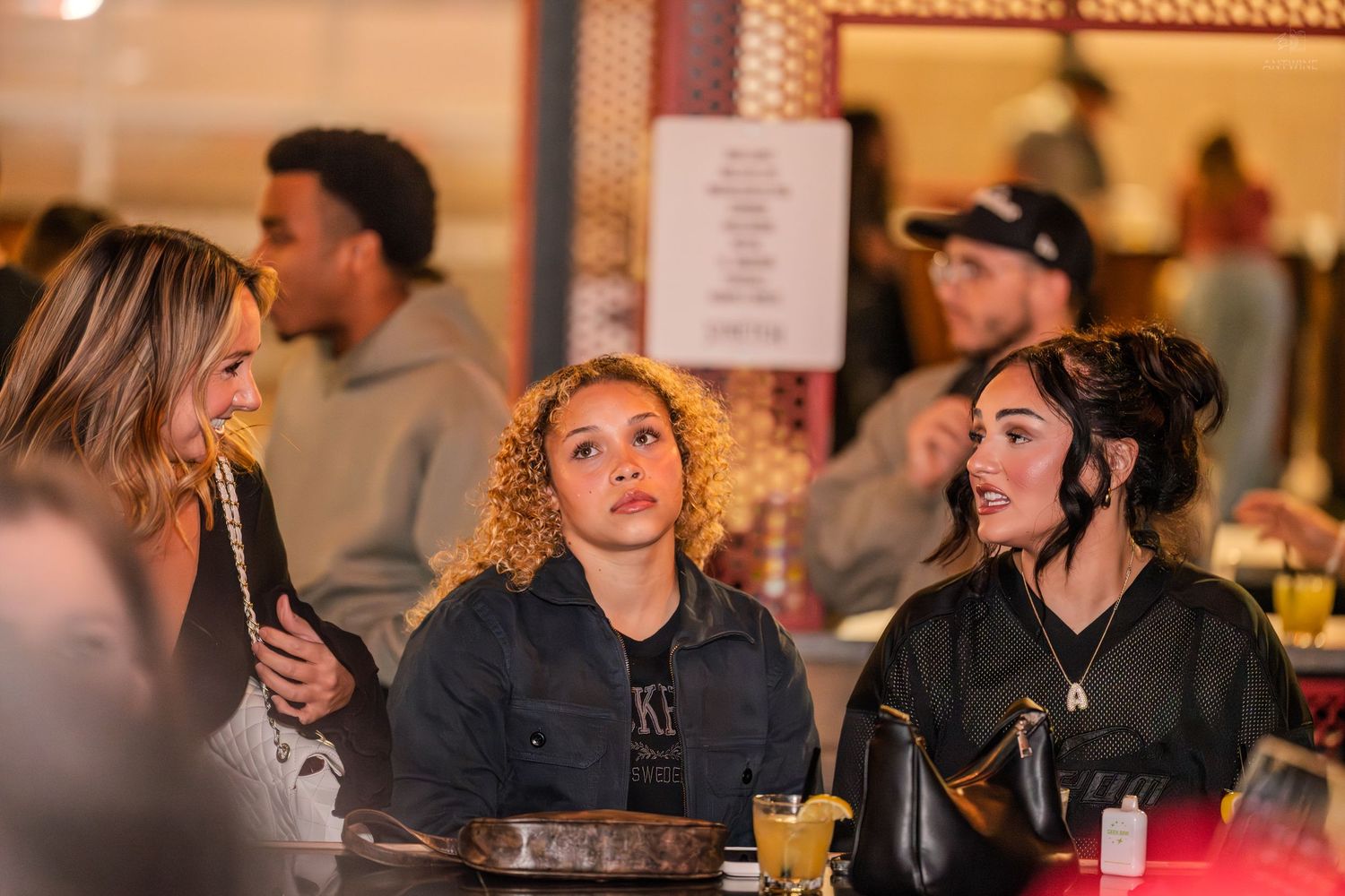 Three young women sitting together at a table in a lively bar or restaurant.