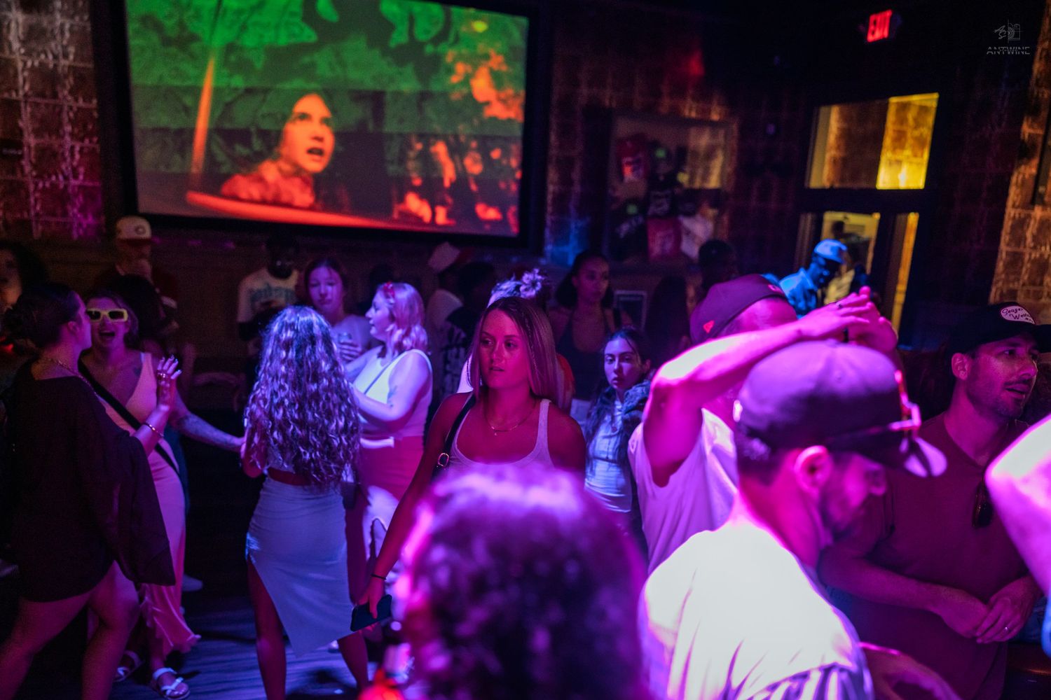 Nightclub dancers under green and purple lights with a large video projection screen above.