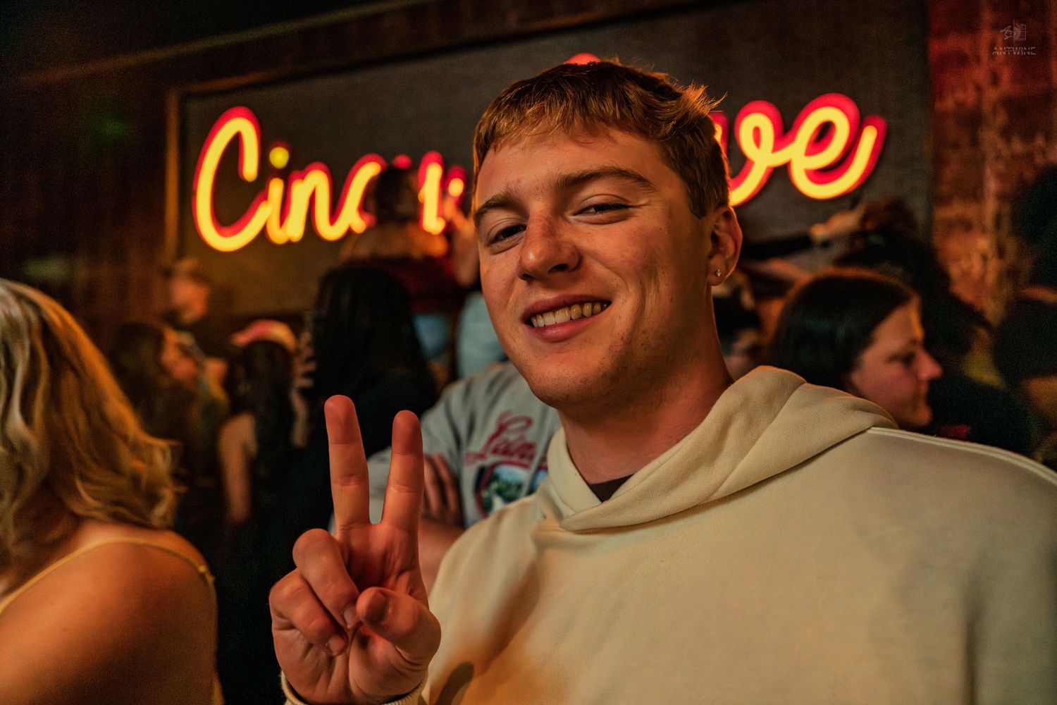 Smiling young man flashing peace sign at a party with colorful neon signs behind him.