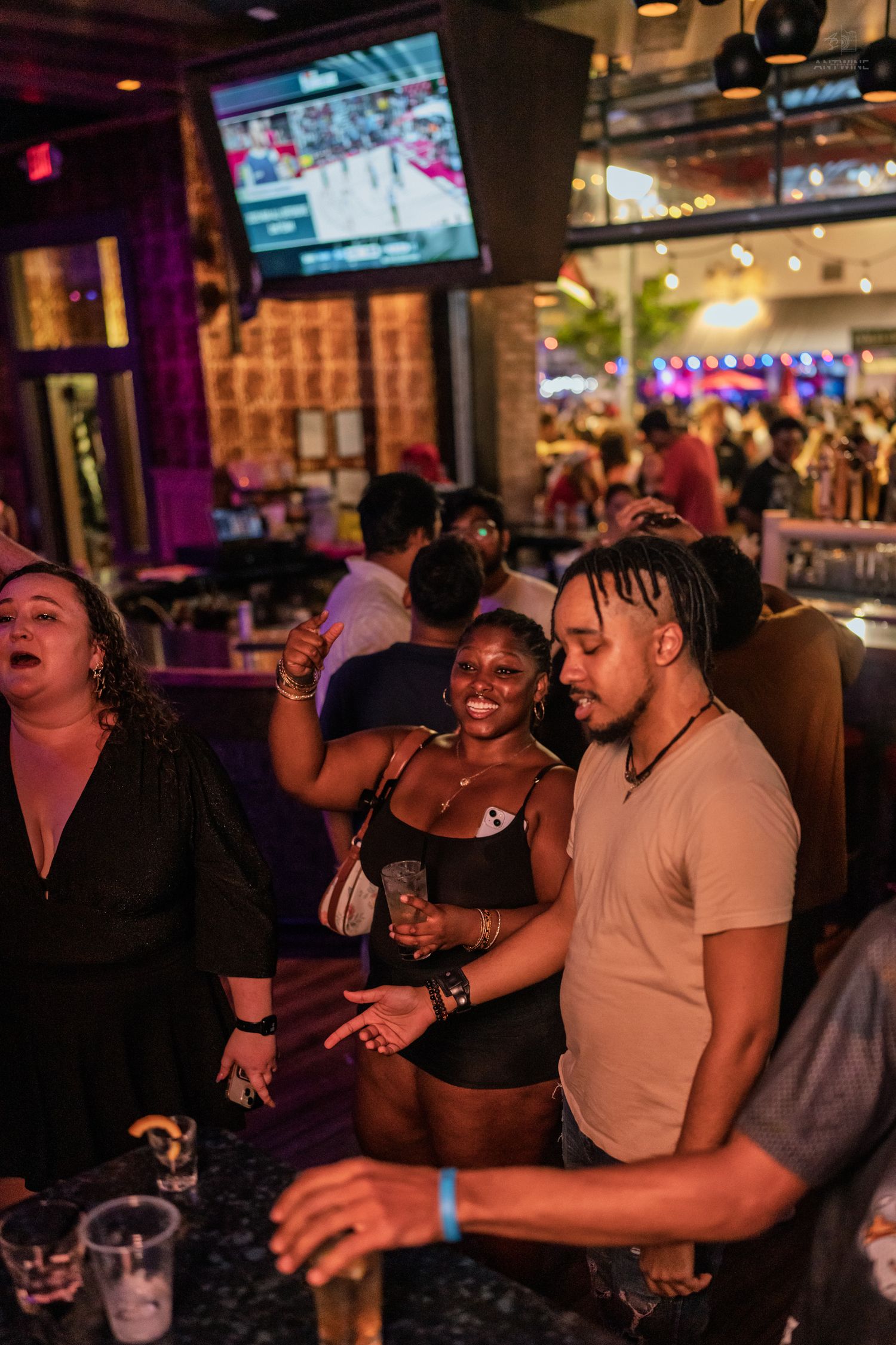 Smiling couple enjoying drinks together at a busy nightclub bar with TV screens above.