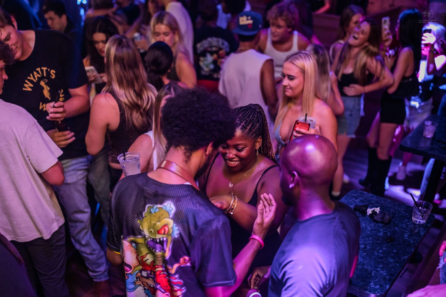 Diverse group of people dancing and socializing under purple lights at a nightclub.