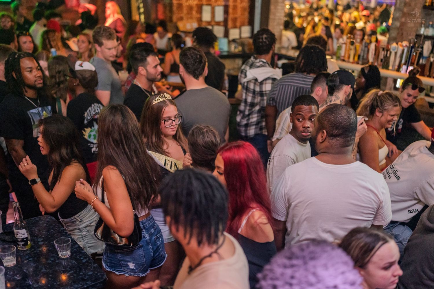 Diverse group of people mingling at a busy bar with colorful lighting and drinks in hand.
