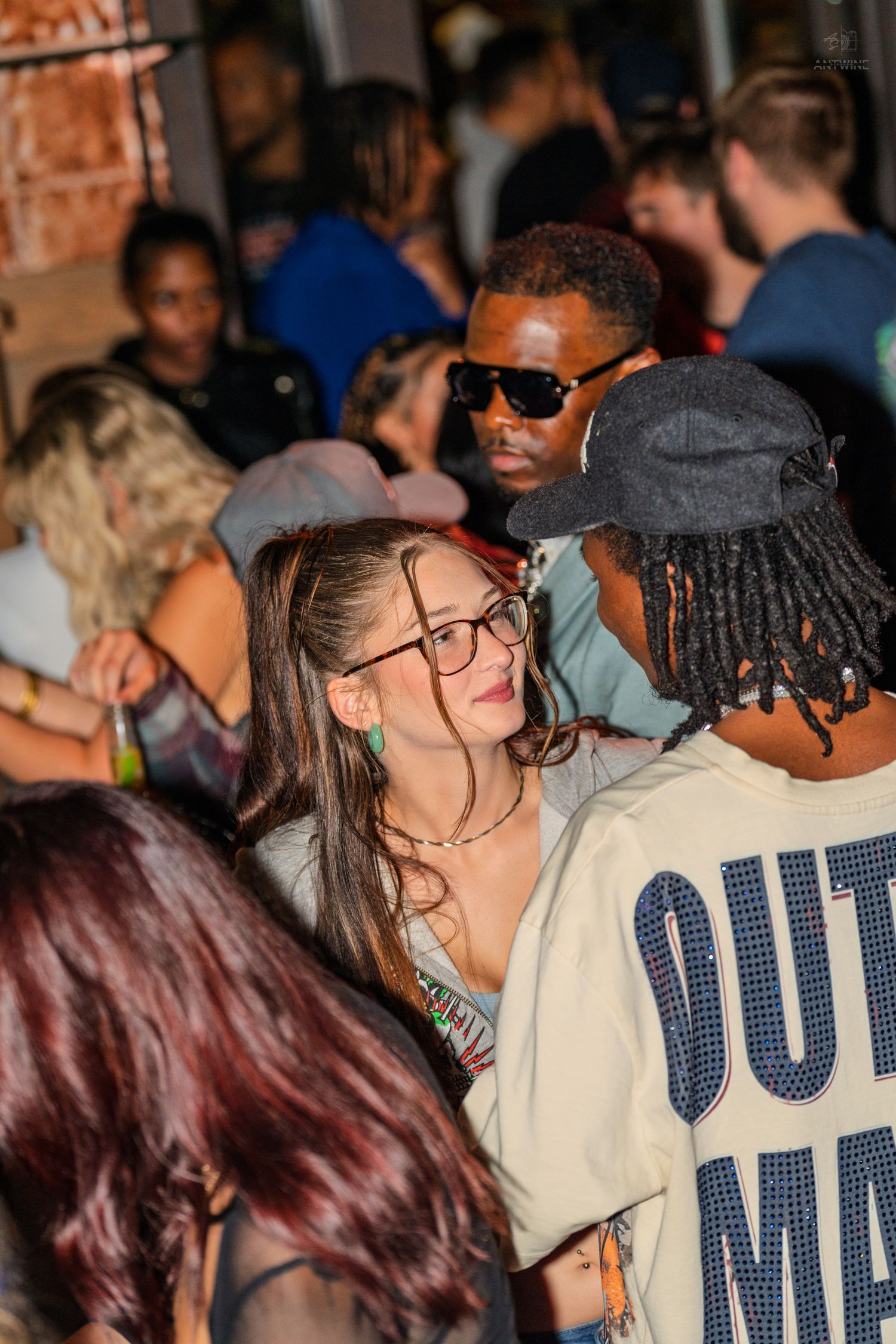 Young woman with glasses chatting with a man in dreadlocks at a lively nightclub event.