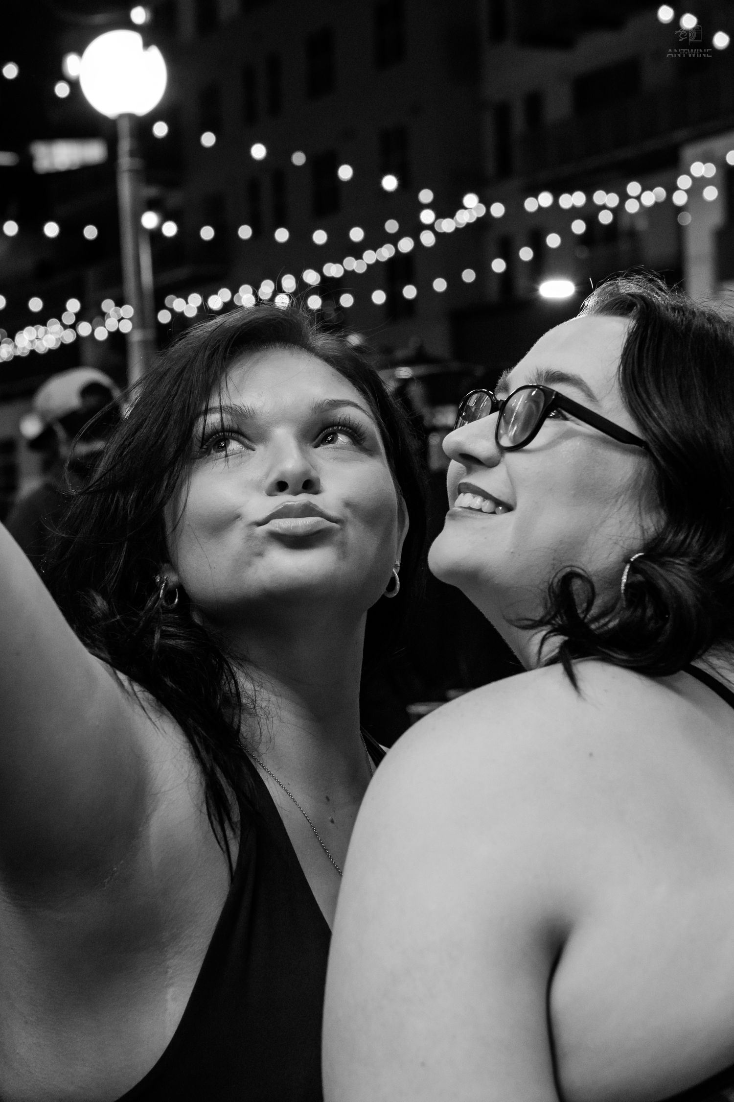 Two brunette women take a selfie outdoors at night under string lights.