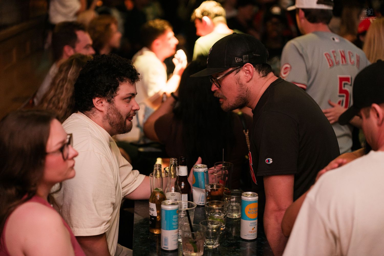 Two men engaged in conversation at a busy bar, with drinks on the table and a lively crowd in the background.