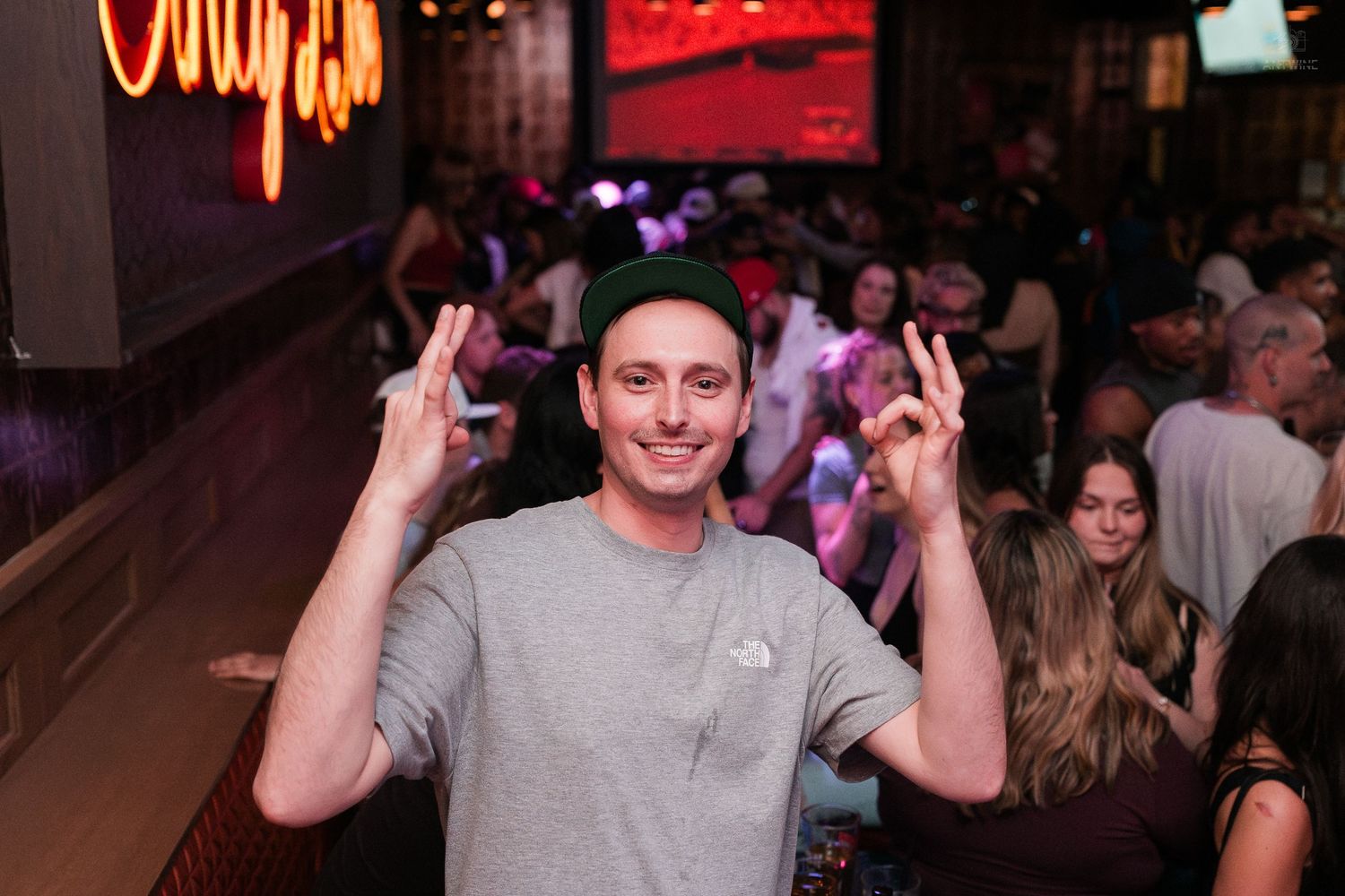 Man in gray shirt and green beanie making peace signs at a crowded bar.