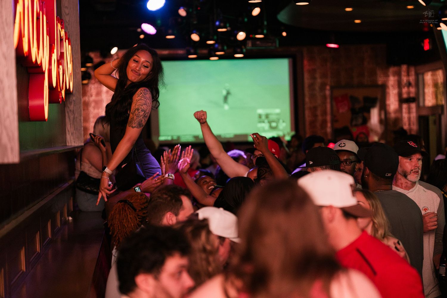 Bartender engages with cheering crowd watching live sports on projection screen at busy bar.