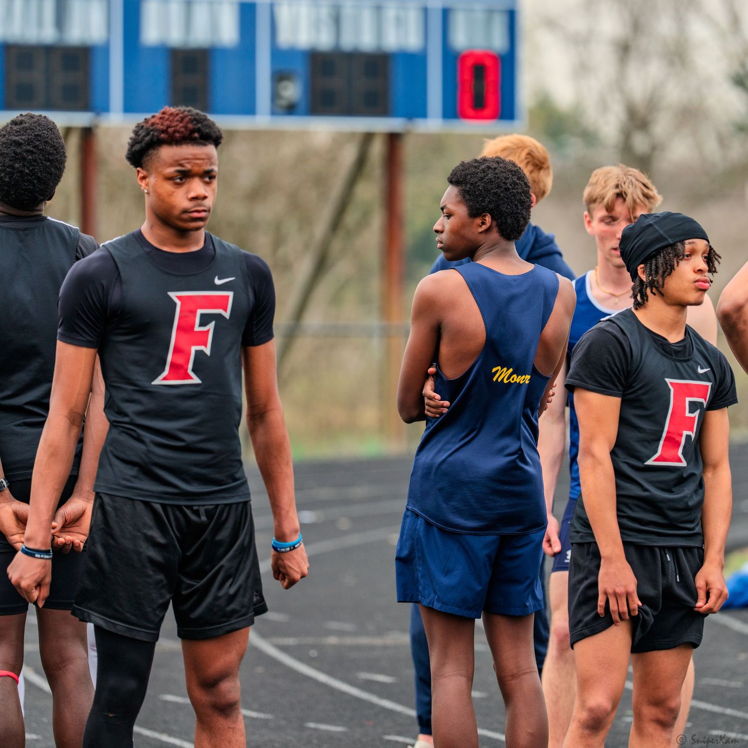 High school track athletes in black F team uniforms waiting near the starting area at meet.