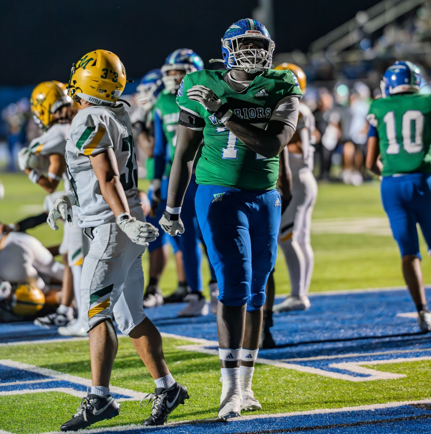 A large football player in green and blue uniform stands on the field during a night game.