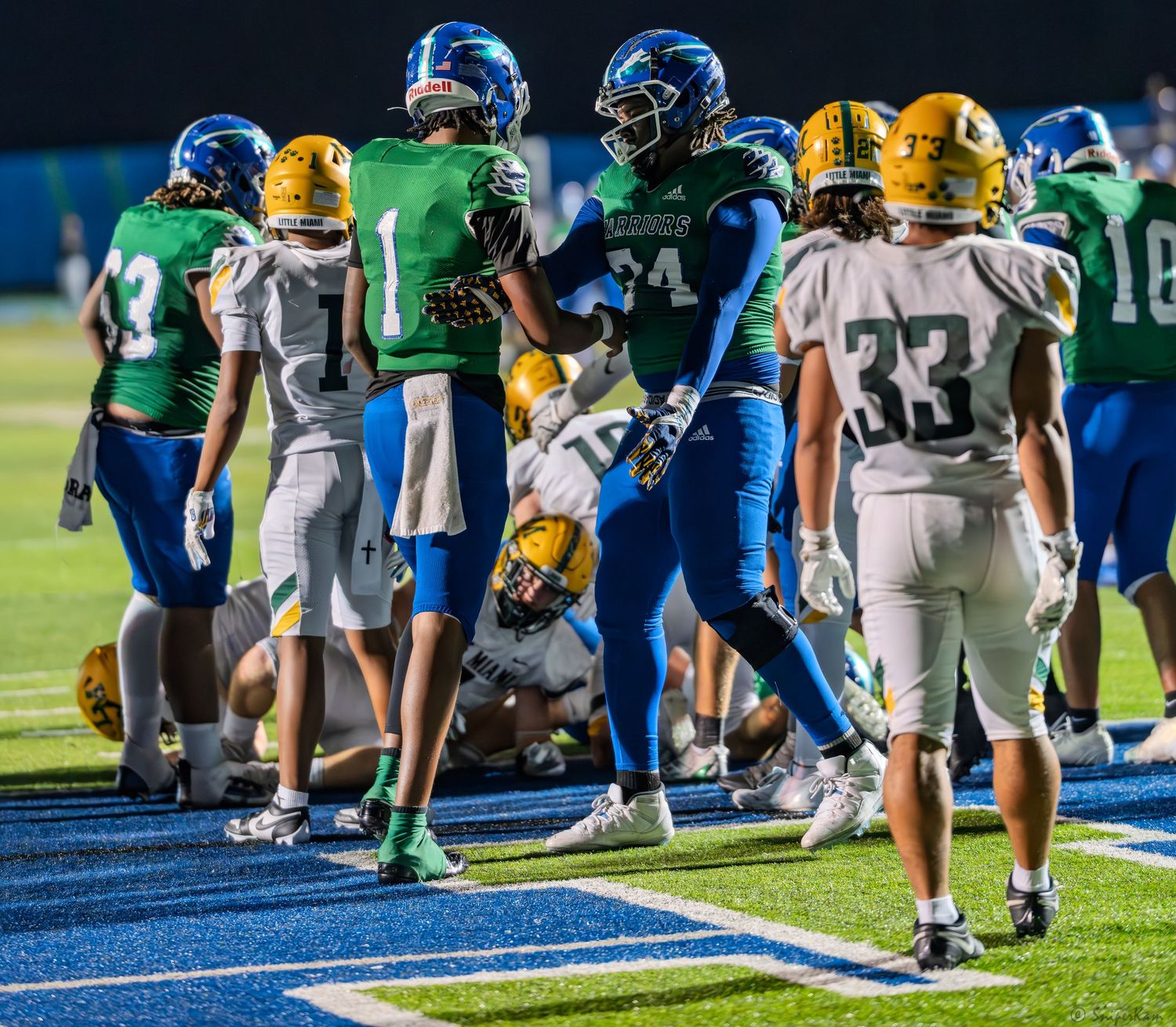 High school football players in blue and green uniforms celebrate after a play during a night game.
