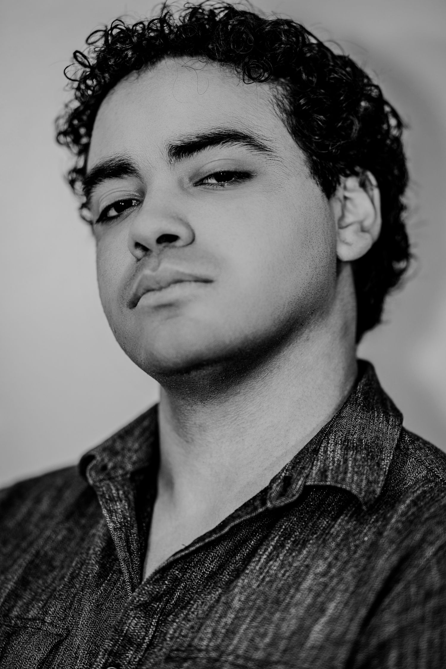 Young man with curly dark hair in a dark shirt looking upward in a moody black and white portrait.