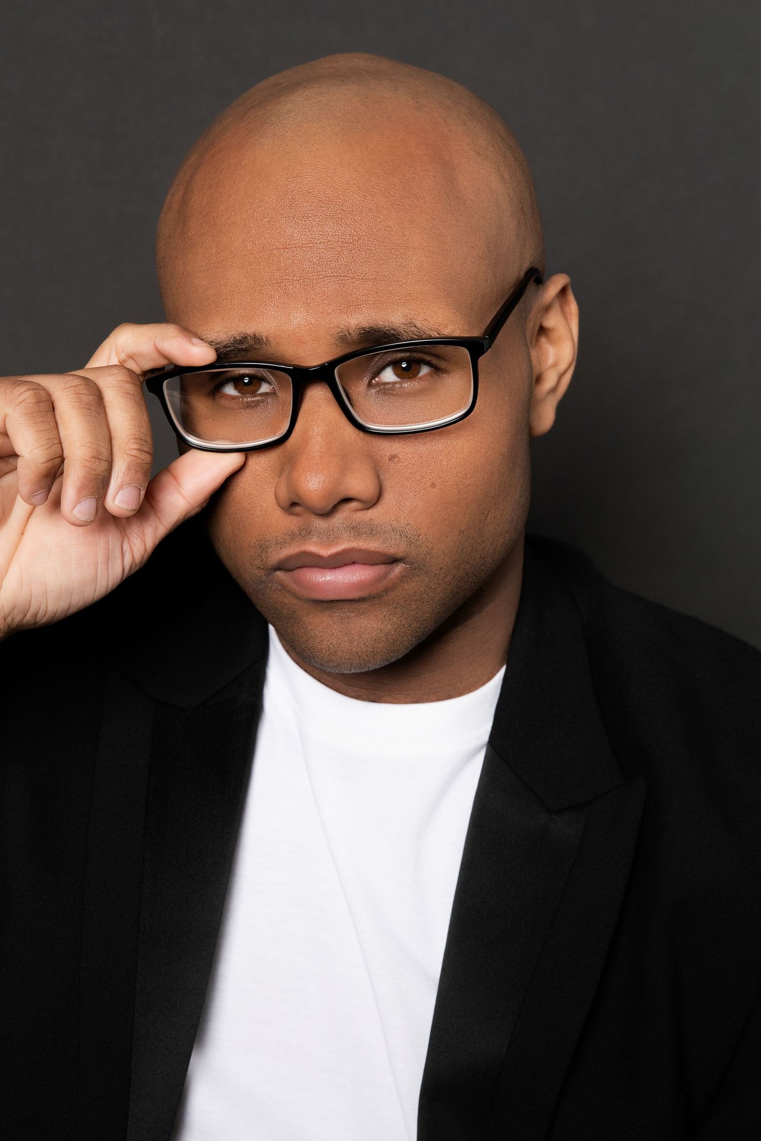 Bald Black man in black blazer holding rectangular glasses, intense gaze in color portrait against dark background.