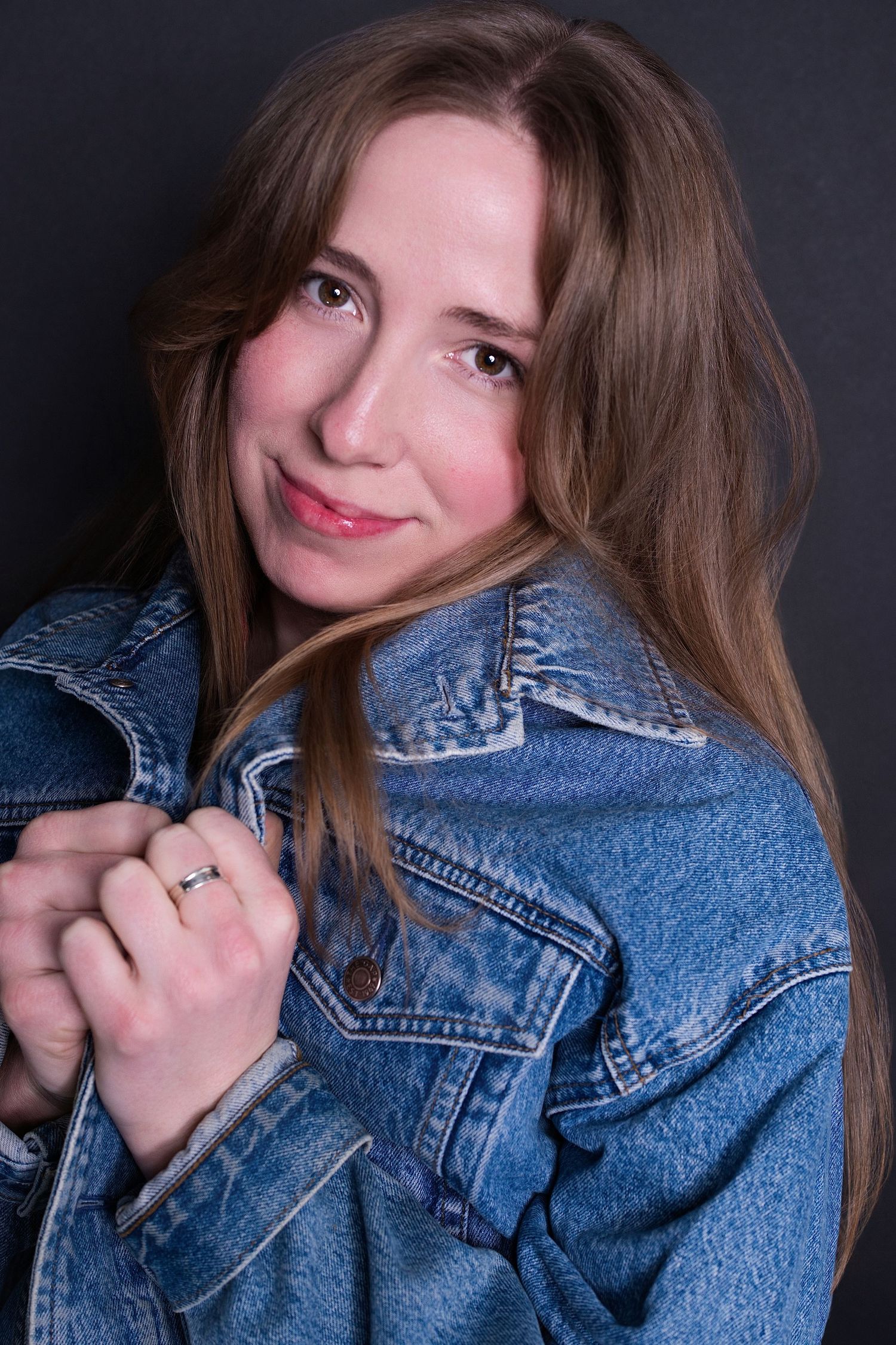 Young woman in blue denim jacket holding lapels, smiling softly at camera against dark background.