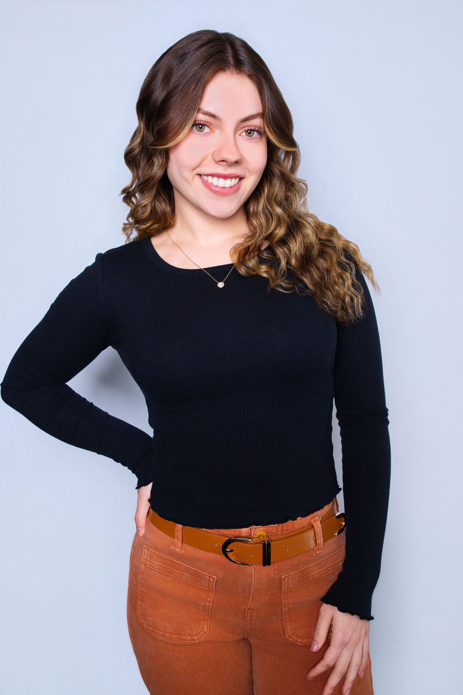 Smiling young woman with curly hair in black long-sleeve top and brown pants poses confidently.