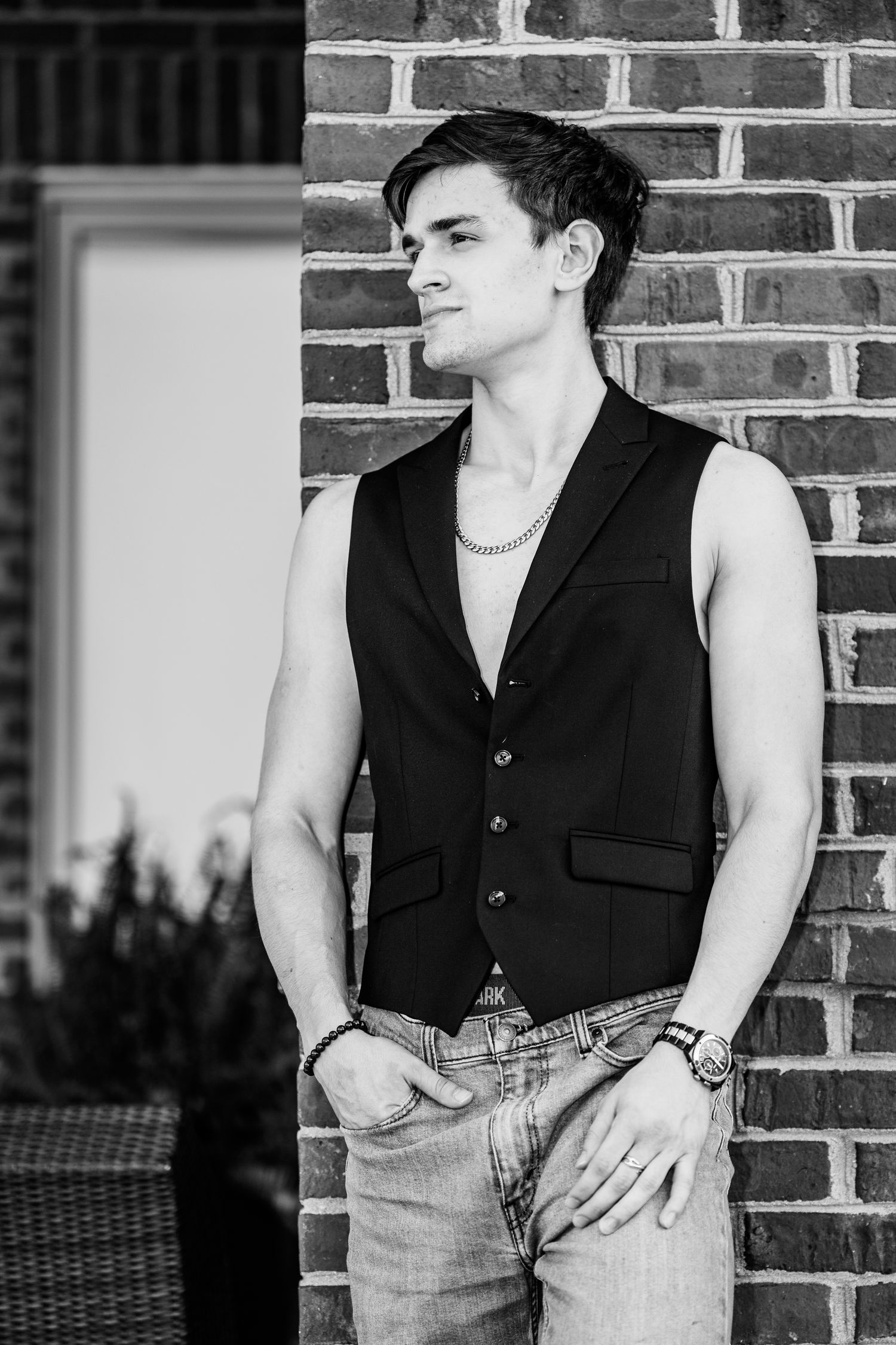 Young man in sleeveless black vest and jeans posing against a brick wall in a black and white photo.