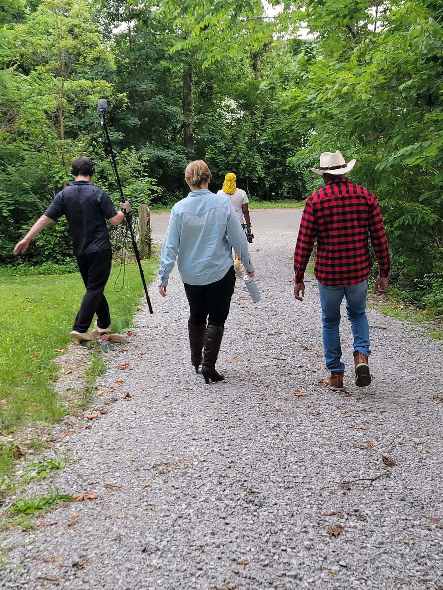 Four people walk down a gravel path through green woods during an outdoor film production.
