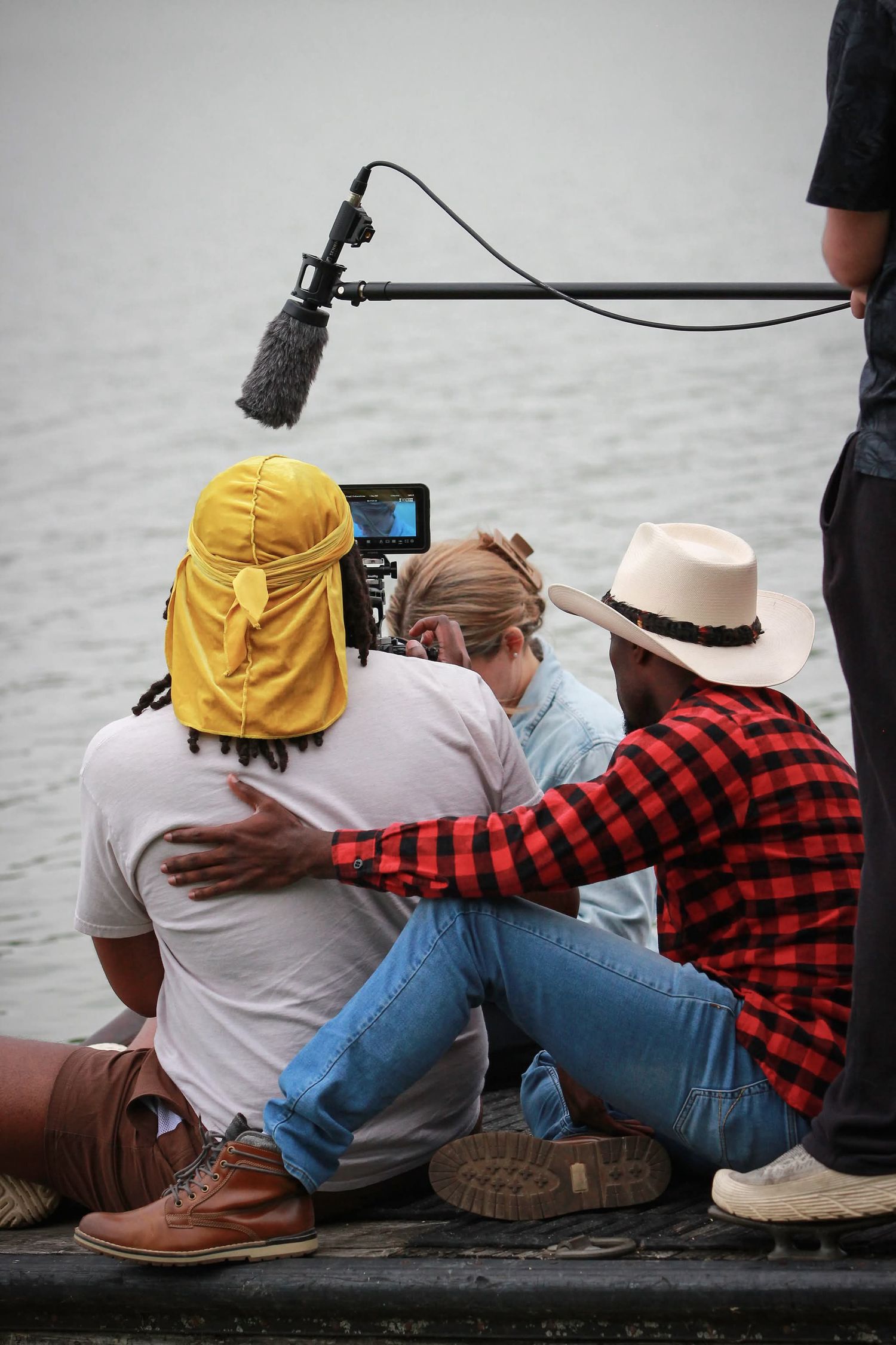 Film crew sits by water with camera and boom mic, two men and woman reviewing footage on set.