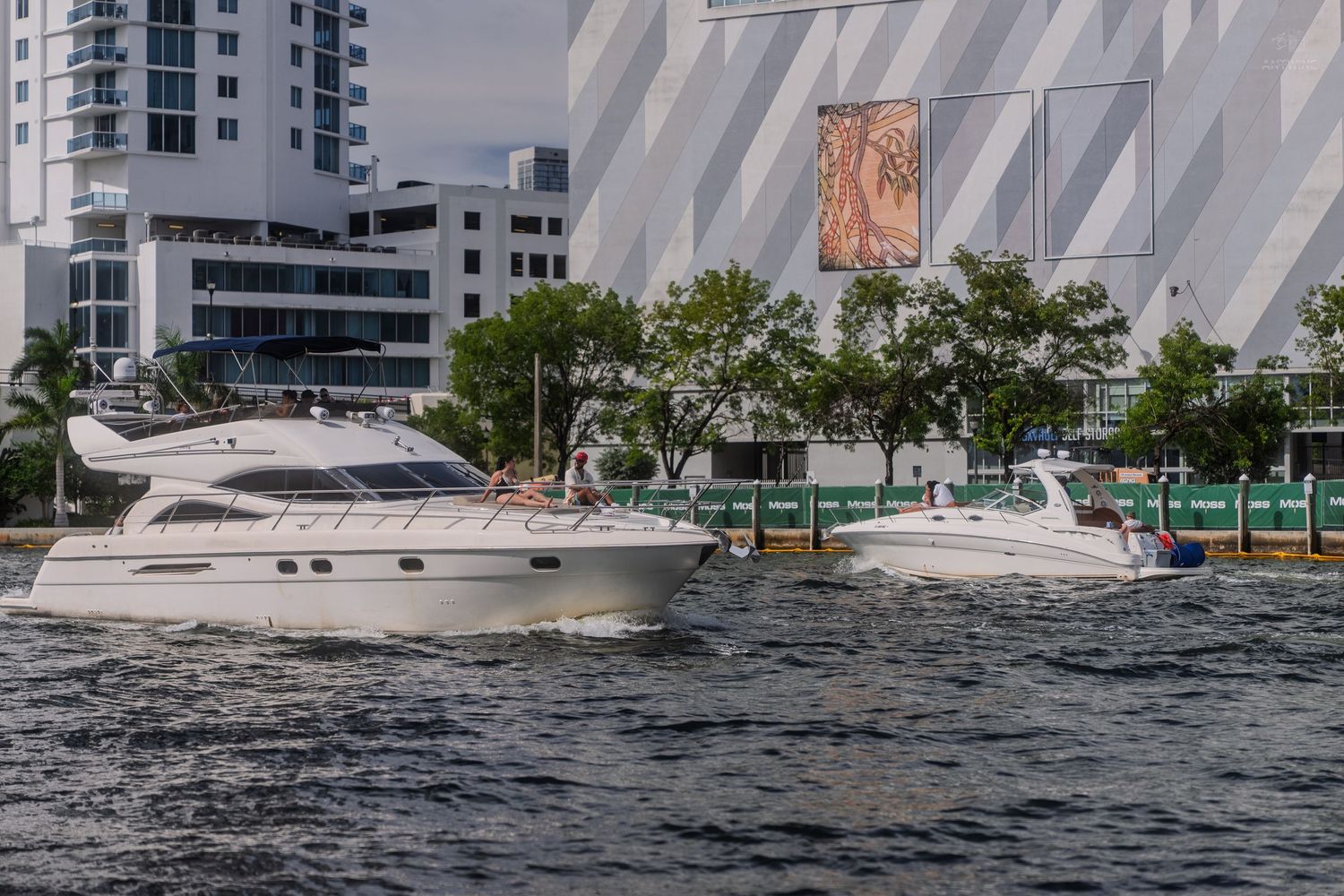 Two luxury white motor yachts cruising along a waterway with modern buildings in the background.