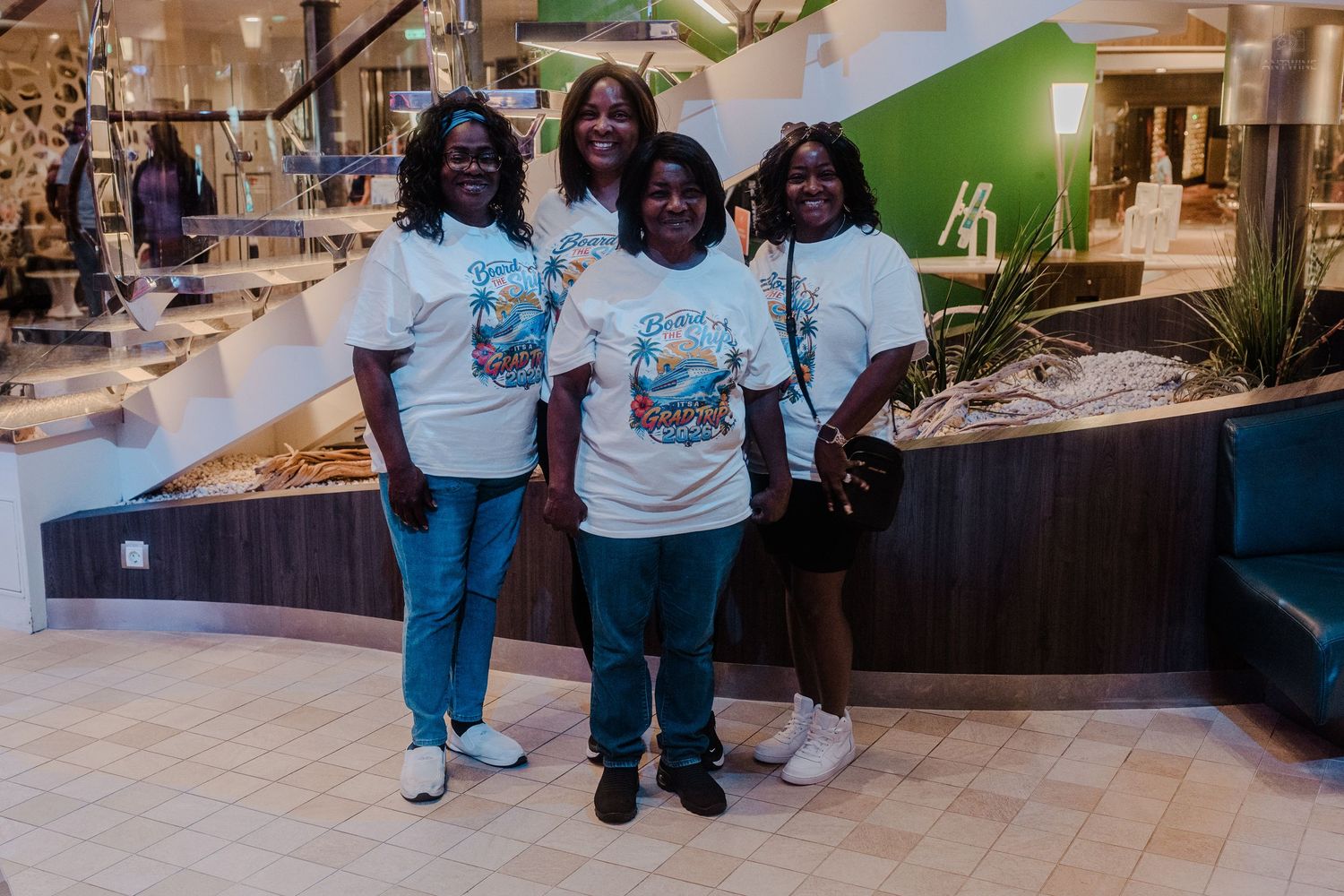 Four women in matching white graphic t-shirts pose together smiling inside a modern cruise ship atrium.