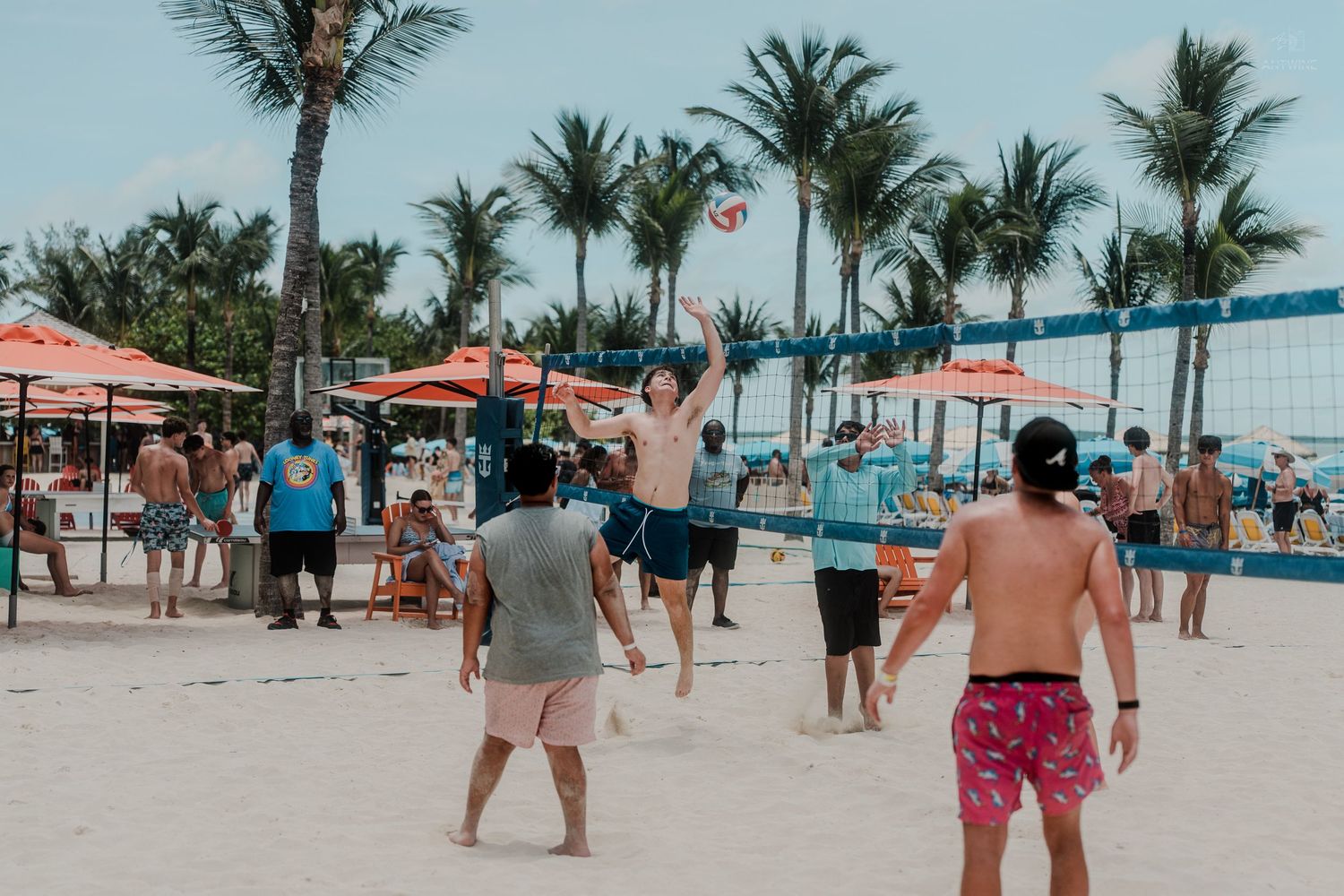 People playing beach volleyball on white sand with palm trees and colorful umbrellas in a tropical resort setting.
