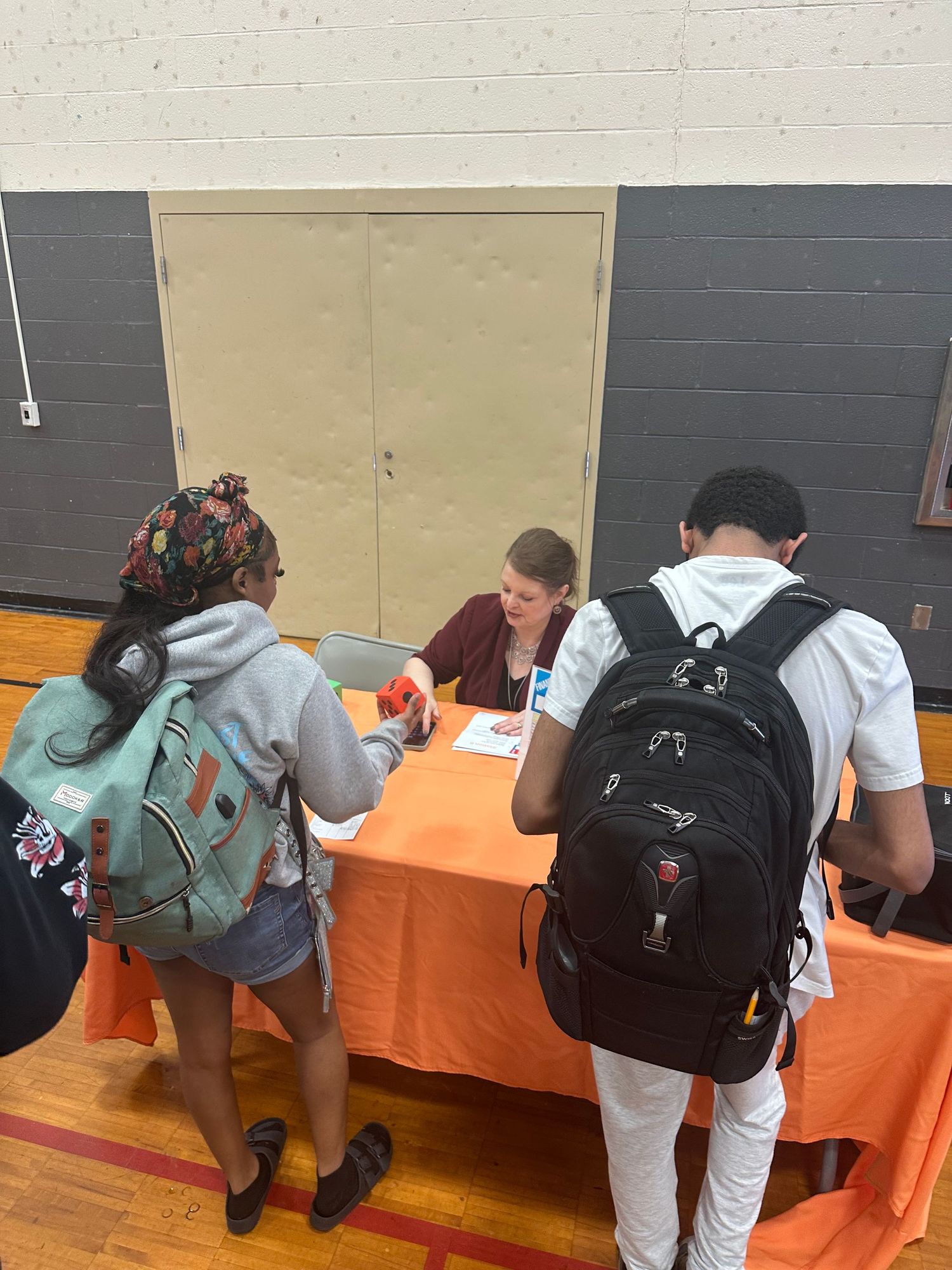 Students with backpacks interact with a representative at an orange-draped table during a school fair in a gymnasium.