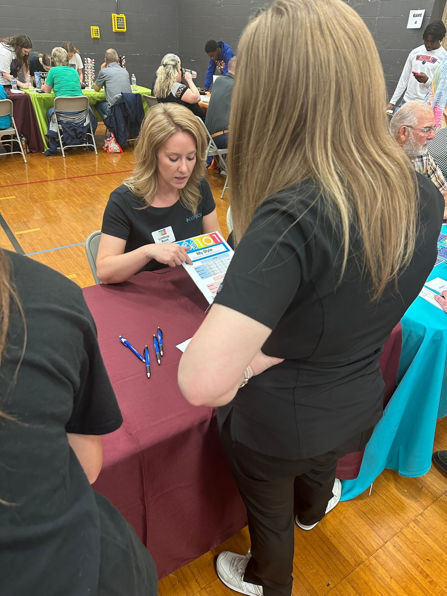 A woman in black presents colorful brochures at a maroon-draped table during an indoor community fair event.
