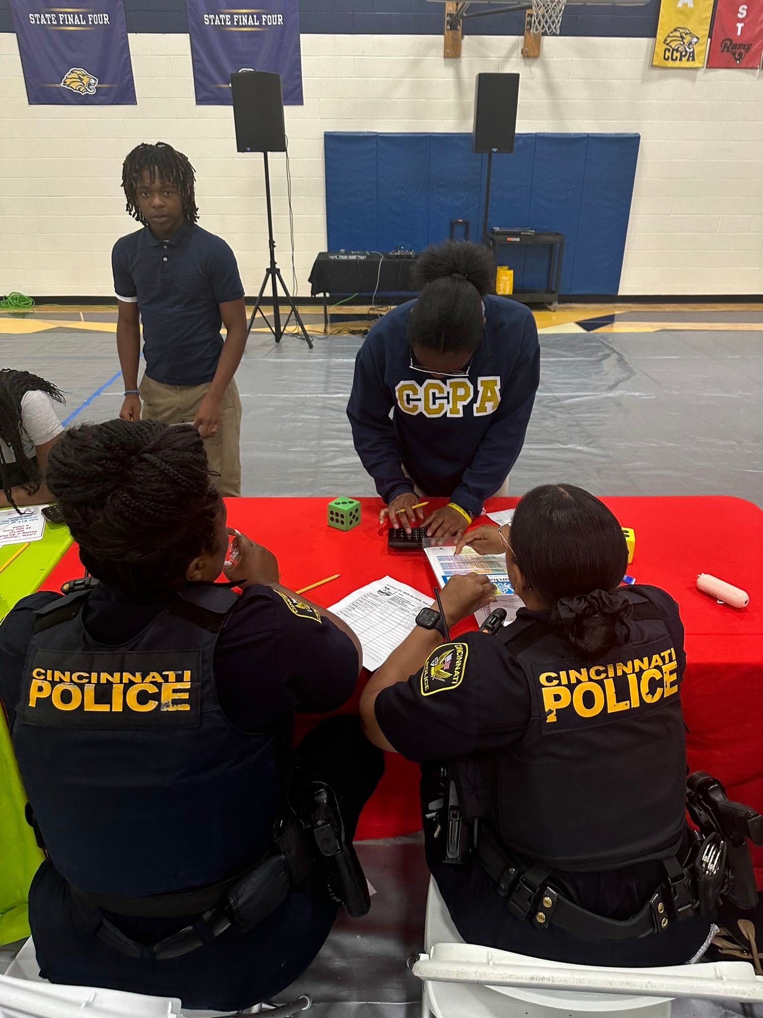 Cincinnati Police officers sit at a red table with students during a school career fair event in a gymnasium.