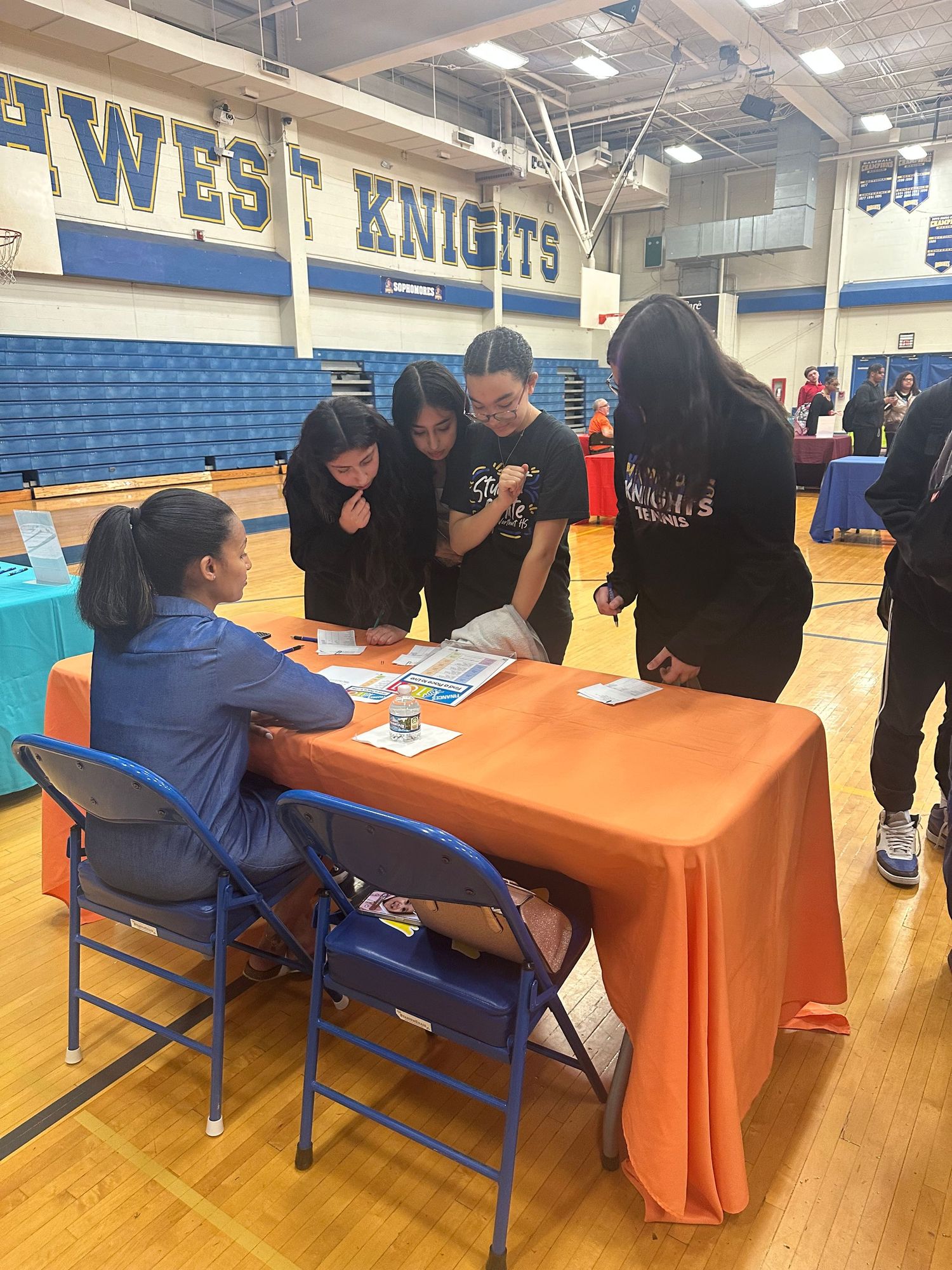 Students gather around an orange table reviewing materials at a school career fair in a gymnasium.