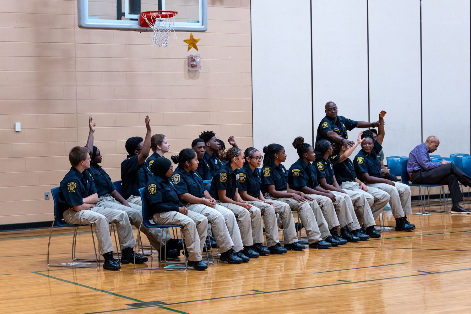 Students in matching uniforms sit in rows of chairs in a gymnasium, with one raising their hand during an assembly.