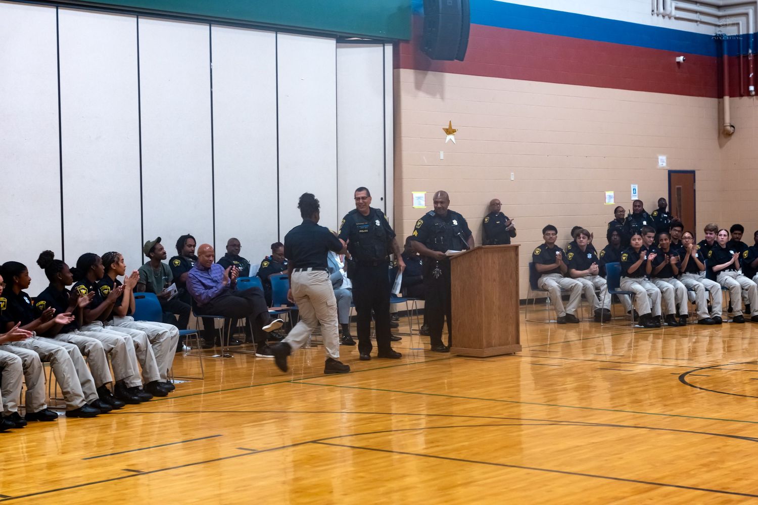 Uniformed students attend a school gymnasium assembly with officials speaking at a wooden podium.