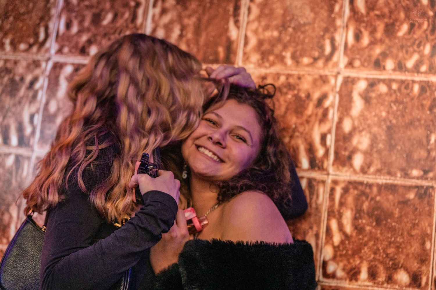 Two women sharing a warm hug near a decorative copper wall at a party.