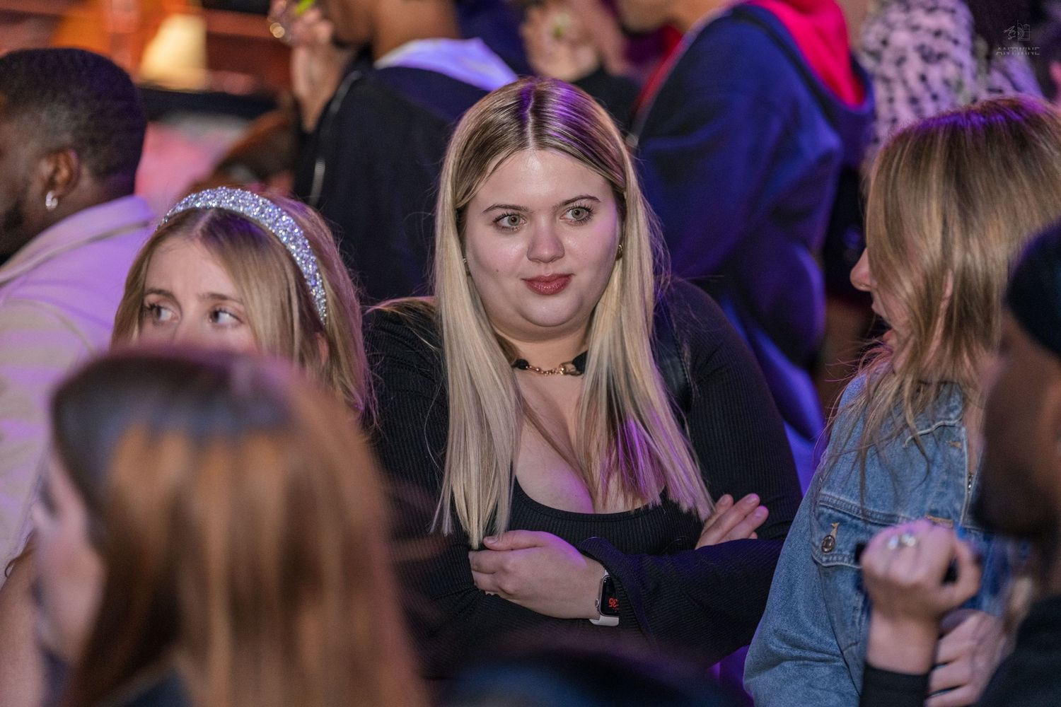 Young blonde woman with arms crossed stands in a crowded concert venue with purple lighting.
