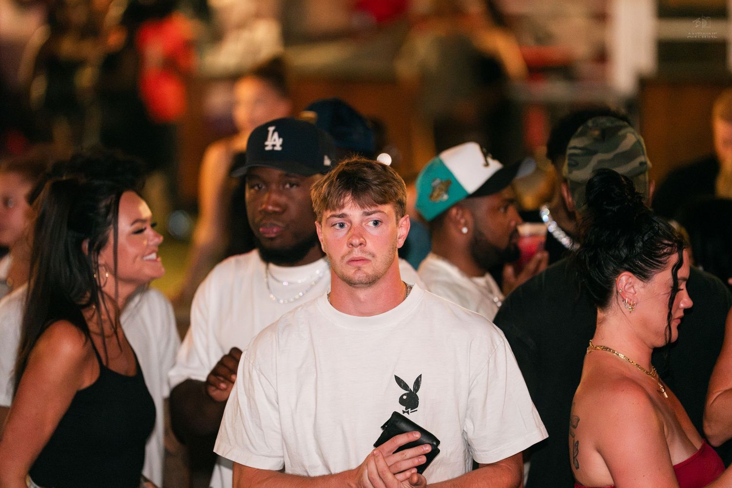 Young man in white Playboy tee holding a drink in a crowded nightclub venue.